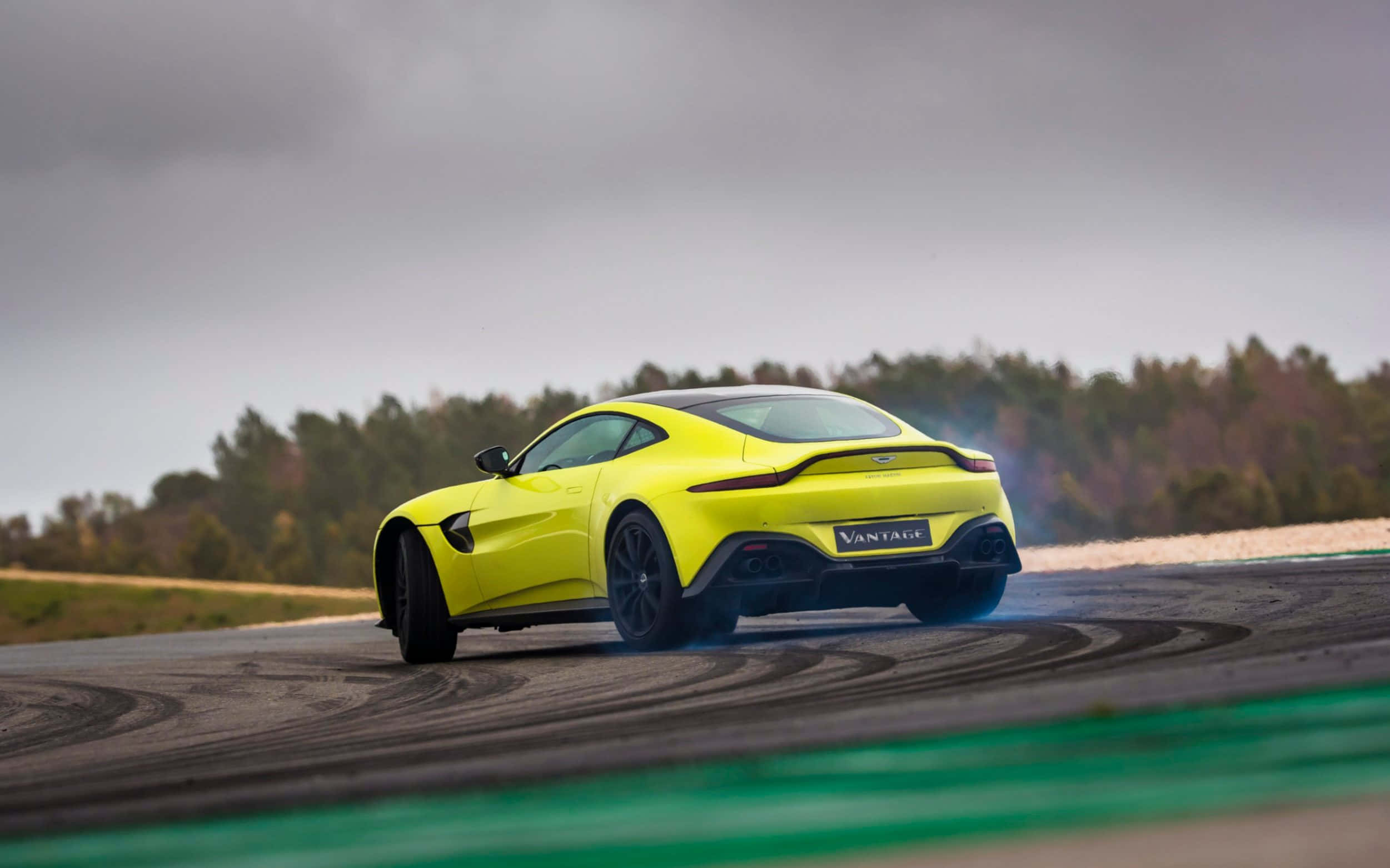 Aston Martin Vantage 桌布 壁紙 — 一輛充滿活力的黃色Aston Martin Vantage在賽道上飄移，留下輪胎煙霧，天空多雲。