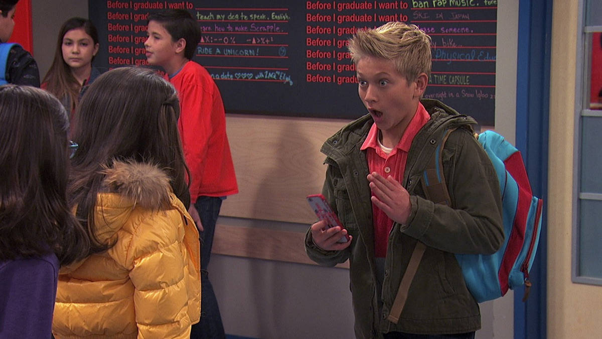 Hudson 驚訝表情 Game Shakers 壁紙 — 一個驚訝的青少年男孩在學校走廊裡拿著電話，同學們聚集在他周圍。