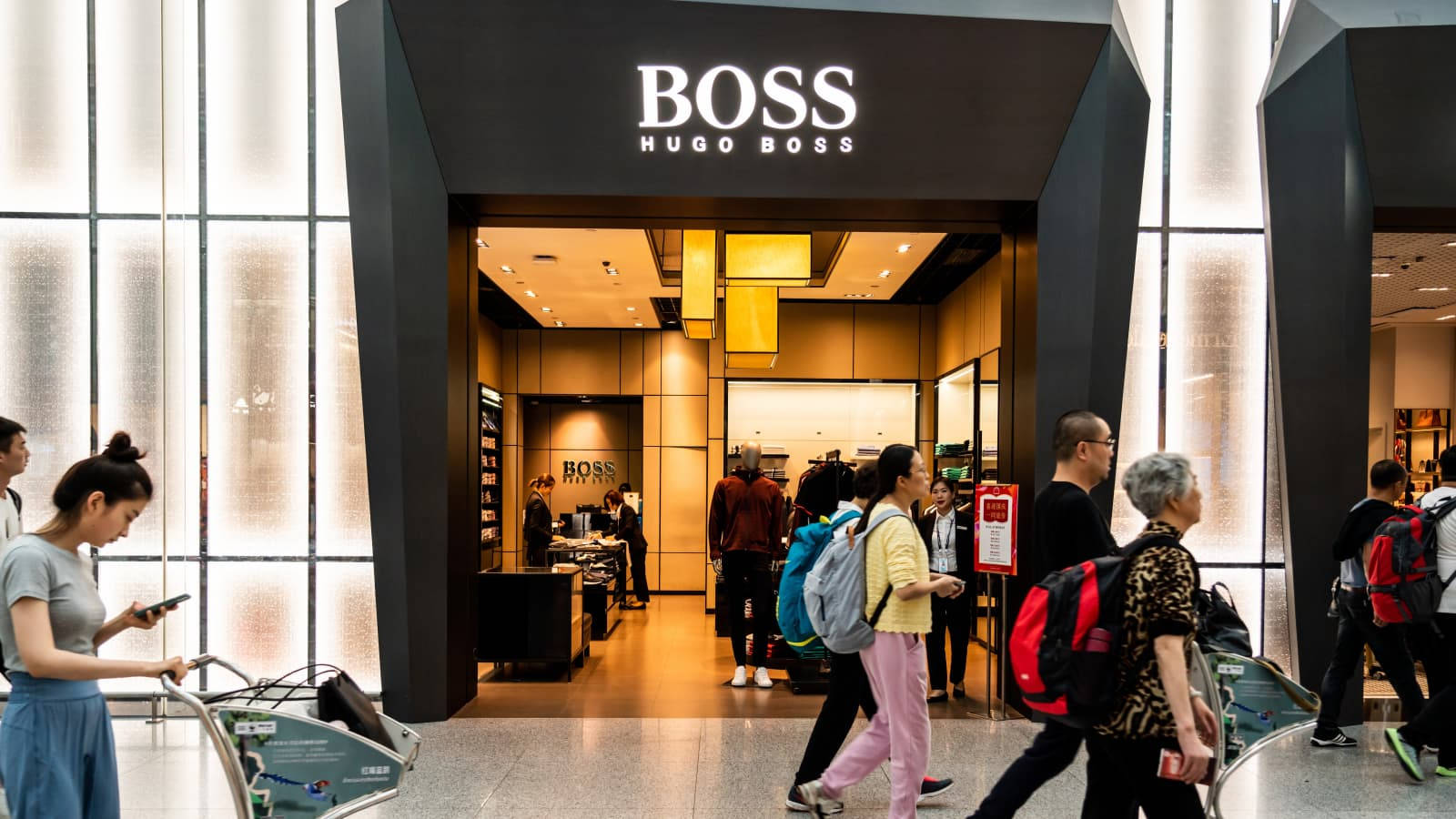 Hugo Boss 機場精品店 壁紙 — 購物者經過一間現代化的Hugo Boss店舖入口，店舖擁有明亮的玻璃牆和溫暖的室內燈光。