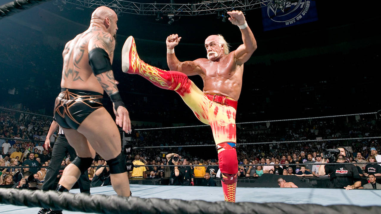 Hulk Hogan 競技場內的戰鬥 壁紙 — 一名穿著紅黃火焰圖案緊身褲的摔角手，在燈光明亮的擂台上，對著咆哮的人群向對手踢出一記高踢。