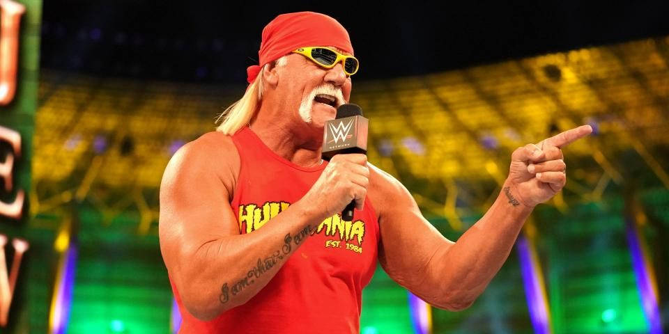 Hulk Hogan 紅色背心造型 壁紙 — 身穿紅色背心和頭巾的霍克·霍根，一邊指著燈火通明的競技場，一邊熱情地對著WWE麥克風說話。