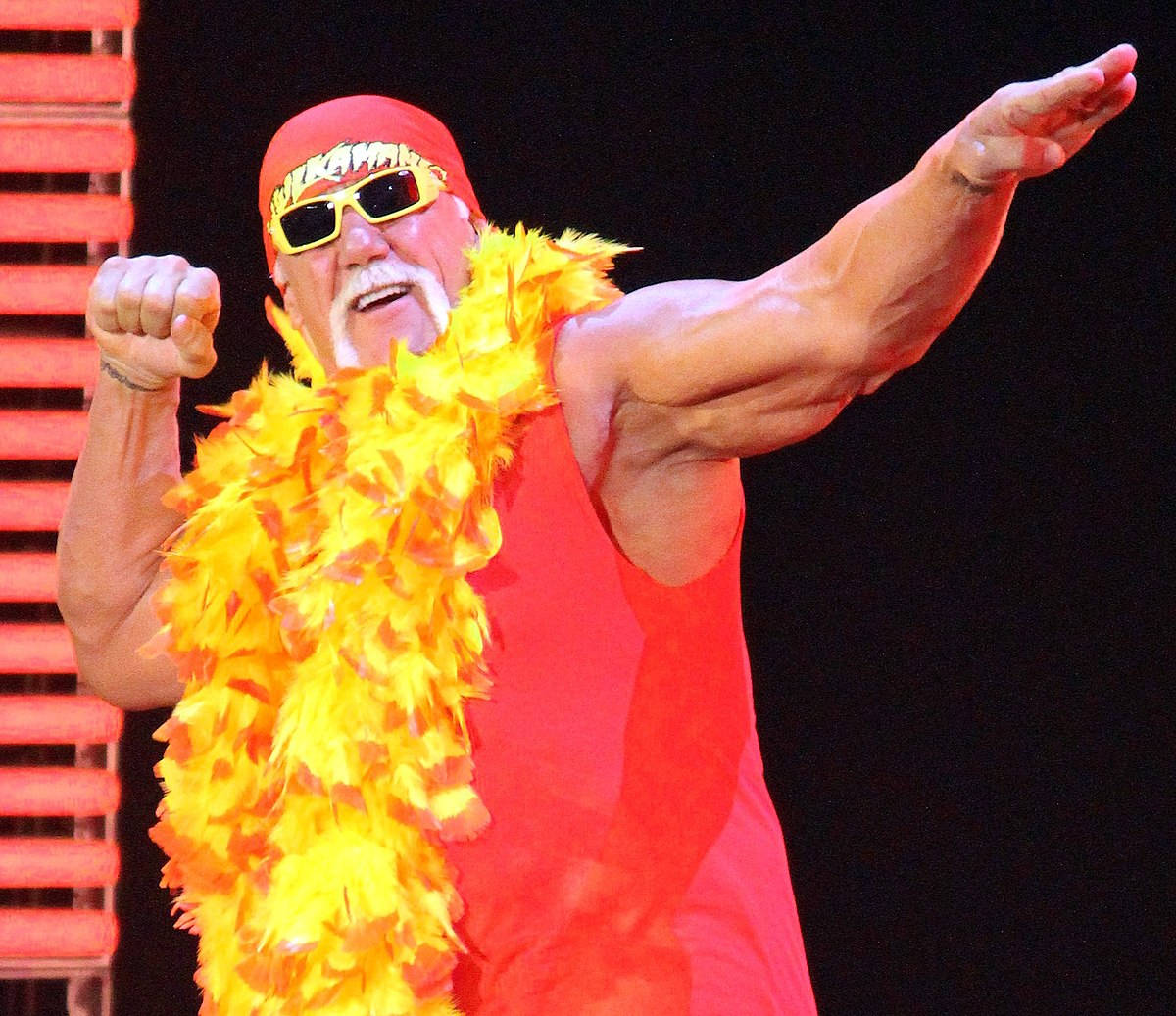 Hulk Hogan 經典招牌姿勢 壁紙 — Hulk Hogan 穿著標誌性的紅色服裝和黃色羽毛圍巾，在明亮的舞台燈光下擺出勝利姿勢。
