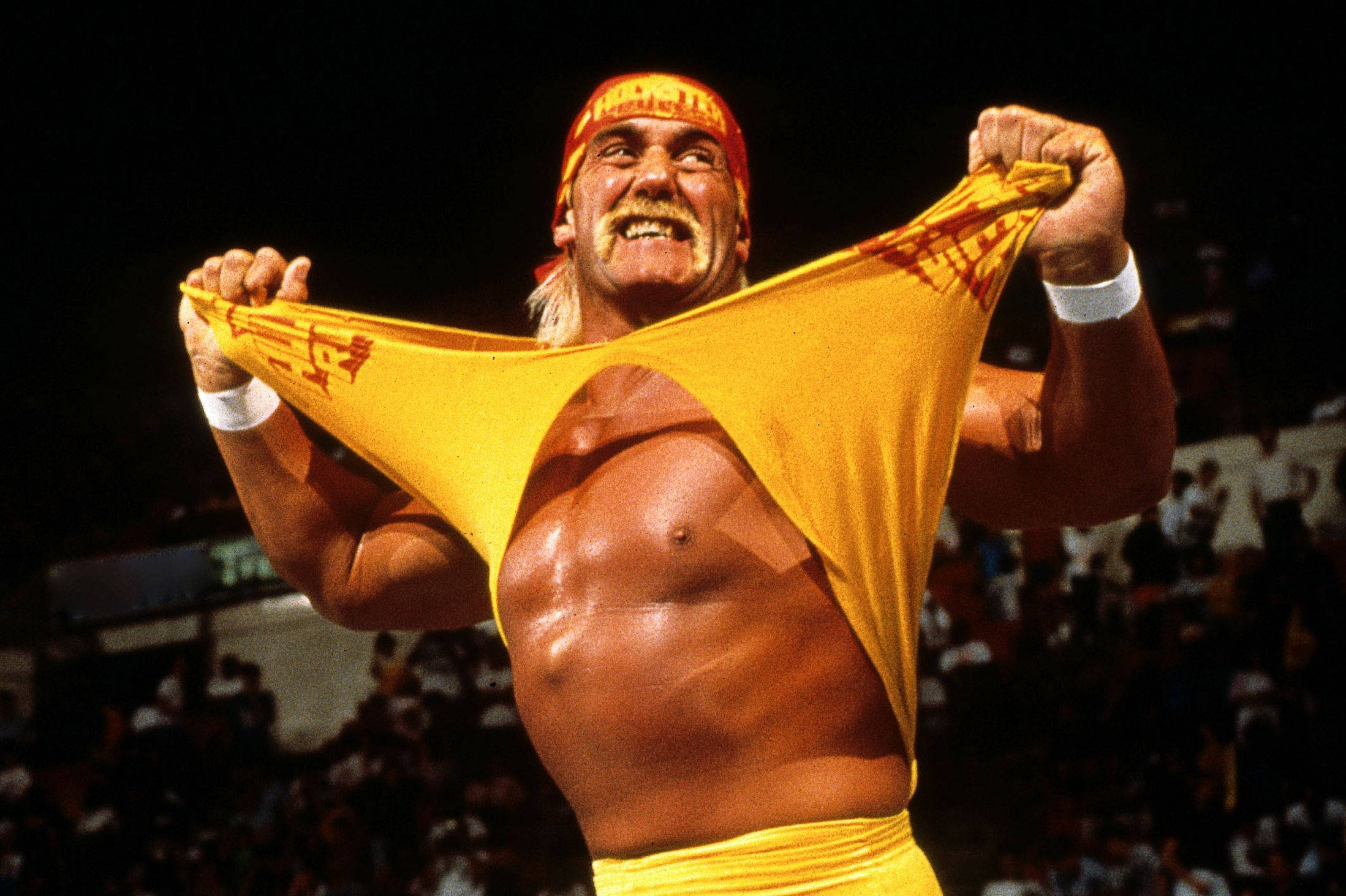 Hulk Hogan 經典黃色 T 恤 壁紙 — Hulk Hogan 在明亮的場館燈光下，以勝利姿態撕破他的黃色摔角衫。