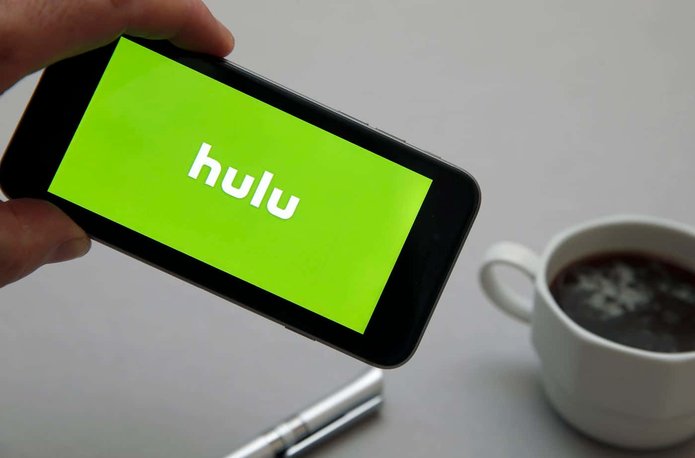 Hulu - 所有串流內容一網打盡 壁紙 — 手裡拿著顯示Hulu標誌的智能手機，旁邊放著一杯咖啡，在簡約風格的桌子上。