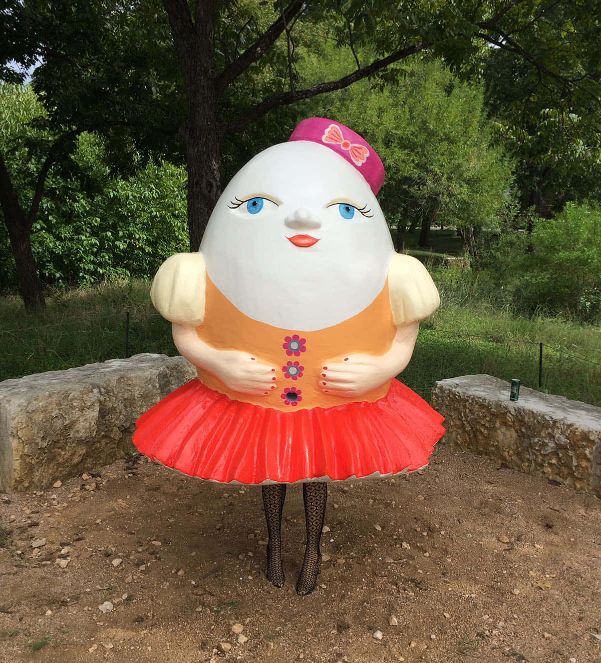 「Humpty Dumpty 坐在牆上的迷人插畫」 壁紙 — 一個巨大的蛋形雕塑，有藍色眼睛和粉紅色帽子，矗立在綠樹成蔭的公園中。