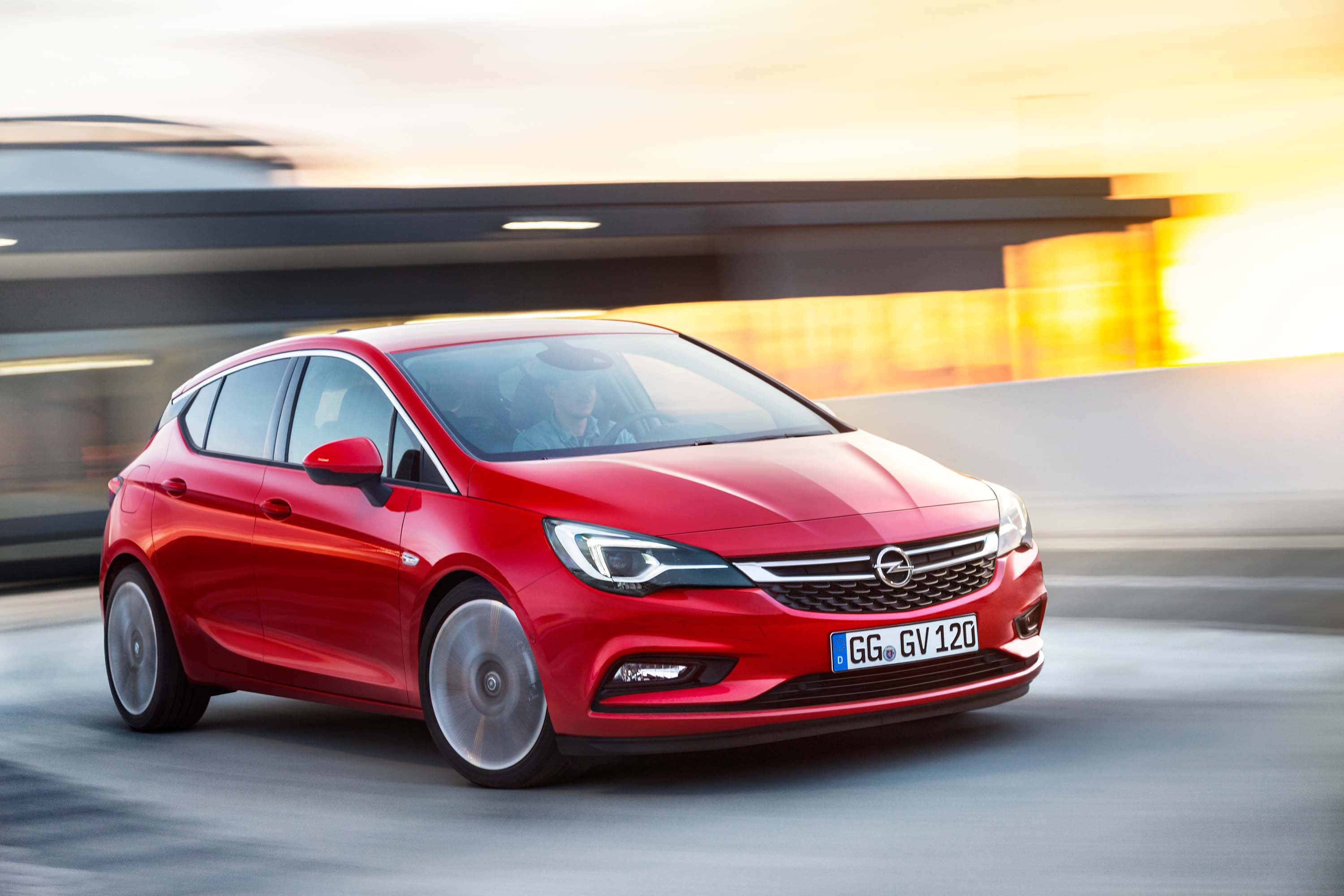 Opel Astra 桌布 壁紙 — 一輛鮮紅色的歐寶雅特掀背車在黃金時段沿現代高速公路疾馳，背景呈現動態模糊效果。