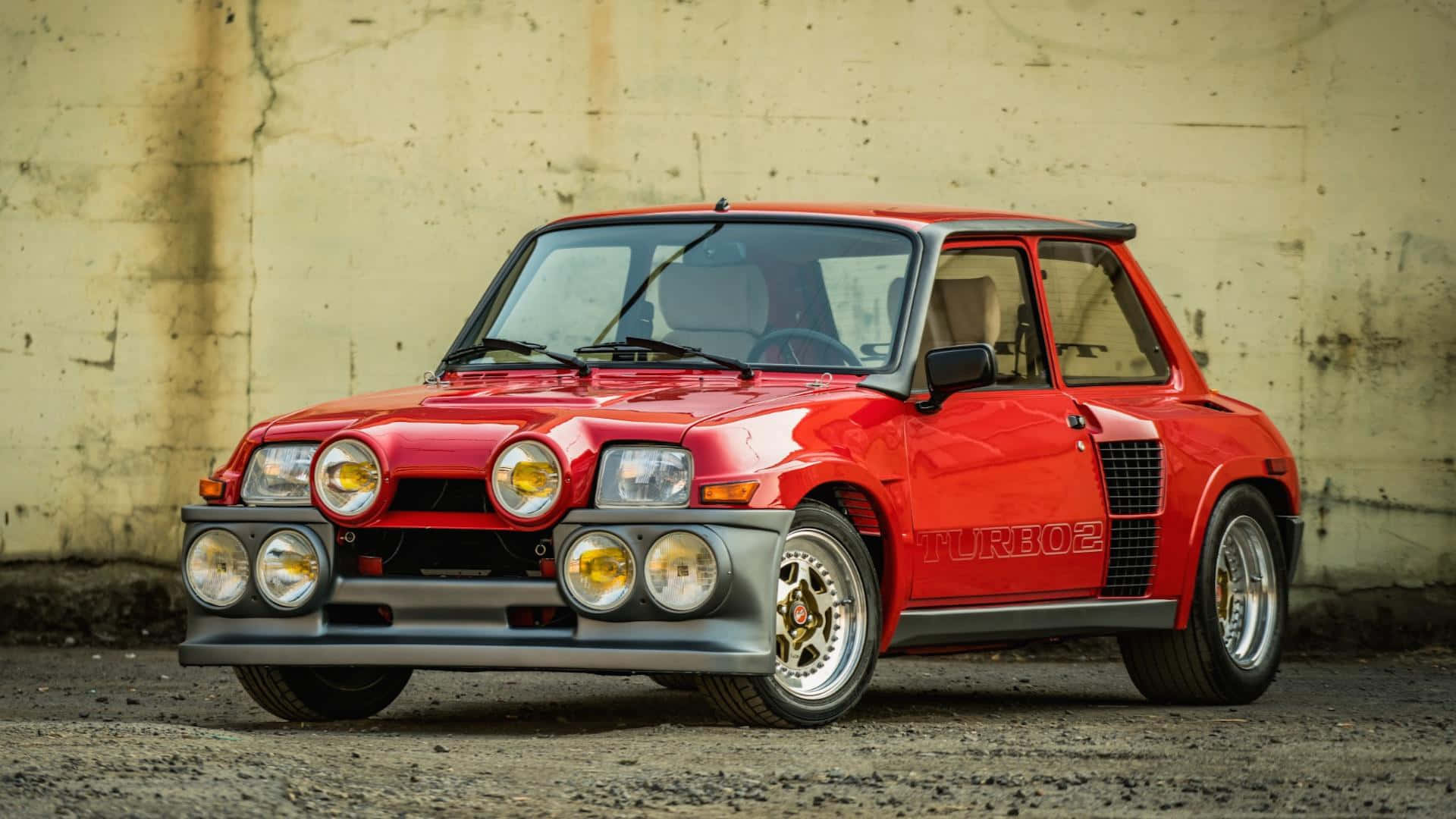 Renault 5 Turbo 桌布 壁紙 — 一輛鮮紅色的雷諾5 Turbo 2跑車，配備六個圓形頭燈和賽車條紋，自豪地停泊在風化的混凝土牆前。