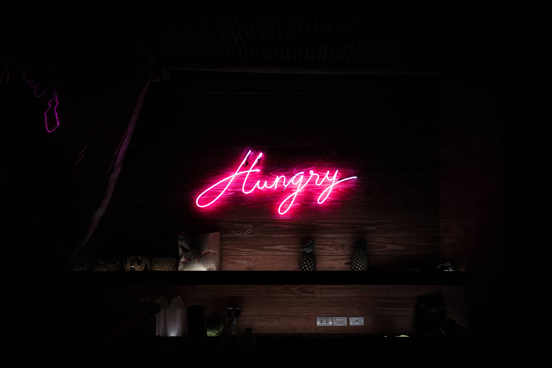 粉紅色霓虹燈牌，上面寫著「Hungry」，位於酒吧架子上方的深色木牆上。