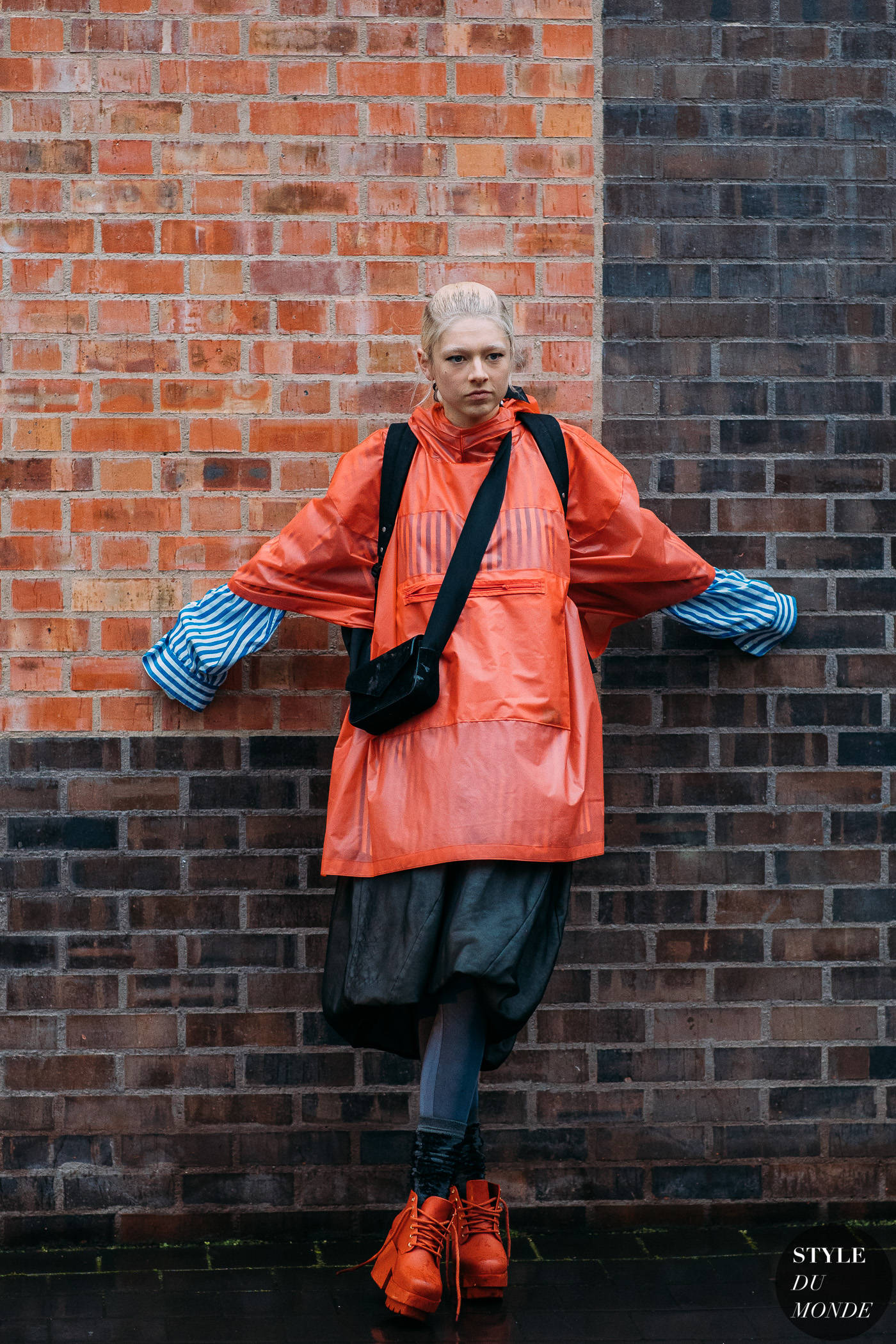 Hunter Schafer 風格造型 壁紙 — 一個人穿著鮮橙色雨衣和條紋袖子，自信地站在風化的磚牆前。