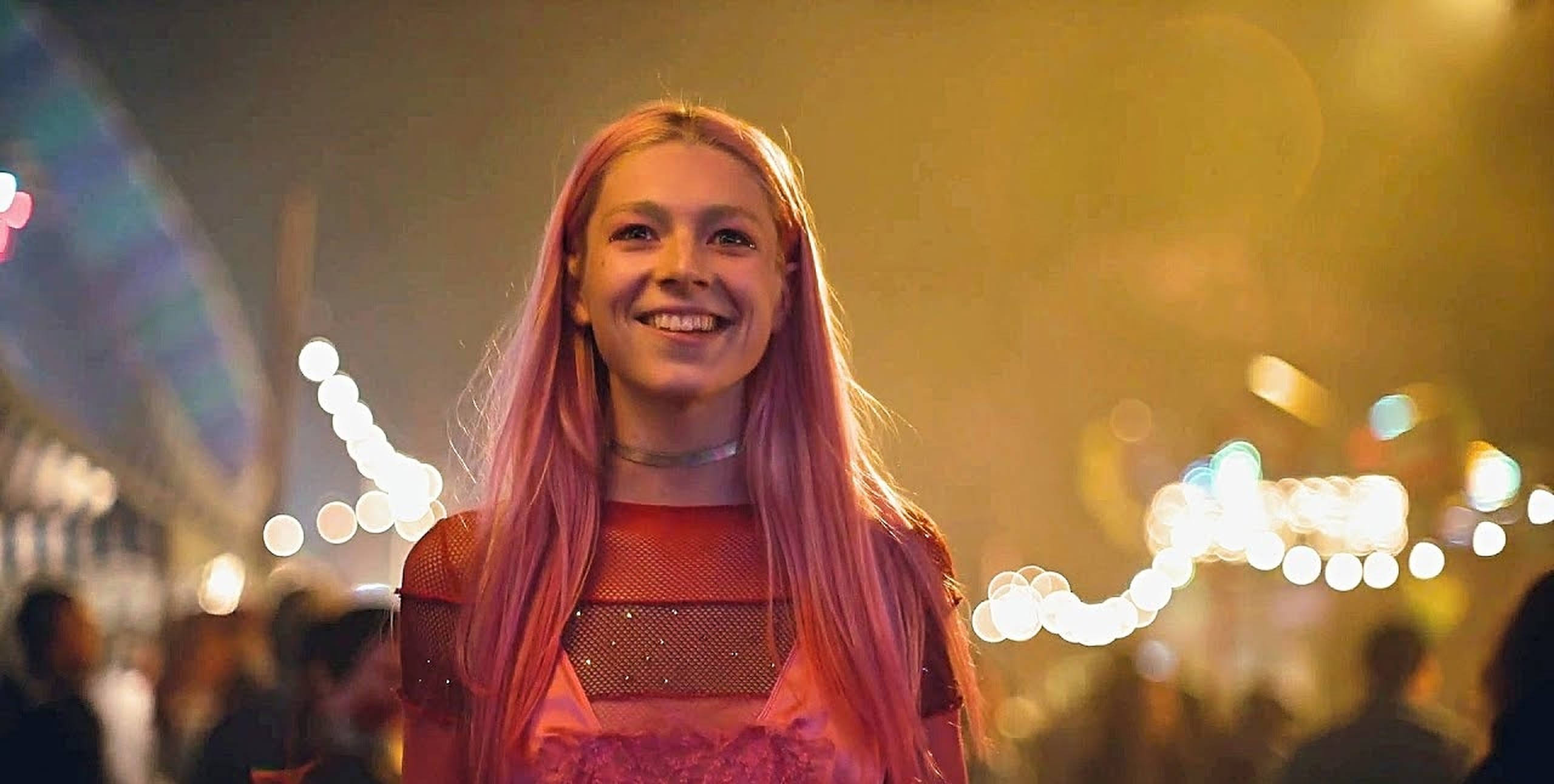 Hunter Schafer Jules 在嘉年華 壁紙 — 一位微笑的年輕女性，粉紅色頭髮，站在充滿活力的夜間嘉年華中，身後有發光的燈光和模糊的人群。