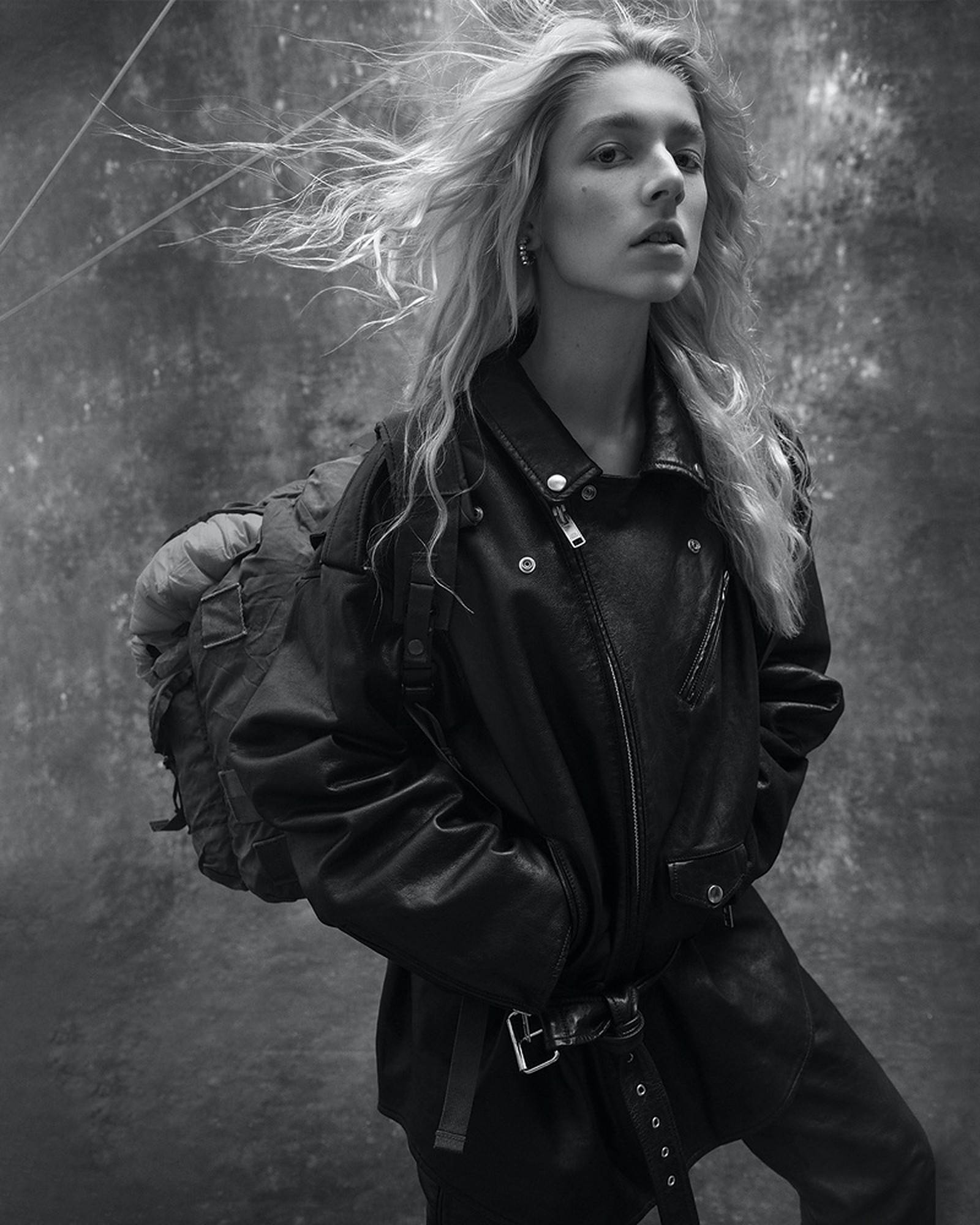 Hunter Schafer 單色寫真 壁紙 — 一位頭髮被風吹拂的女性，穿著皮夾克和背包，在紋理灰色背景前以單色呈現。