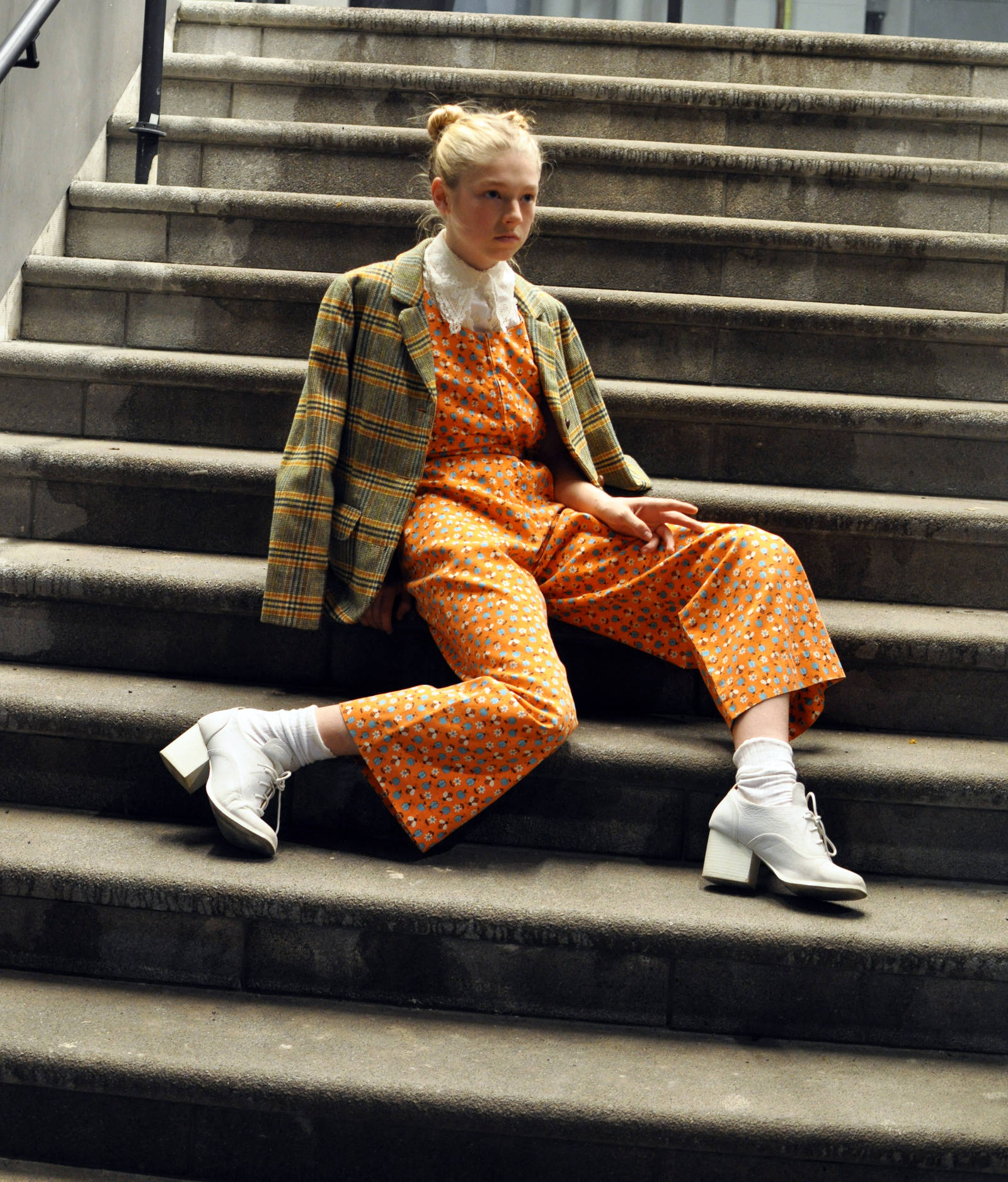 Hunter Schafer 模特造型 壁紙 — 一位年輕女性身穿大膽的橙色圖案連身褲和格子西裝外套，從容地坐在風化的石階上。