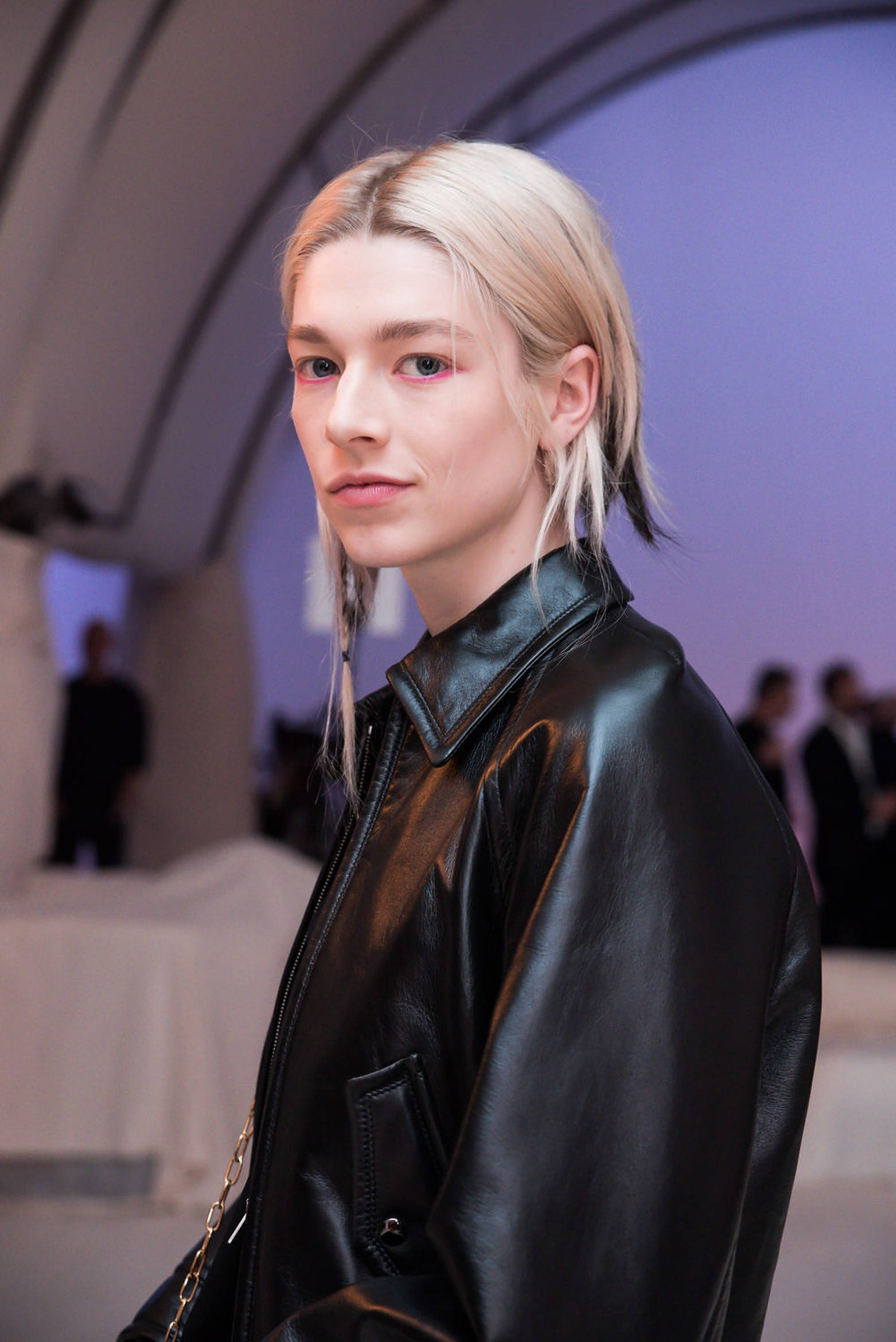 Hunter Schafer 皮衣造型 壁紙 — 一位金髮模特兒穿著黑色皮褸，在柔和的紫色環境燈光下平靜地凝視鏡頭。