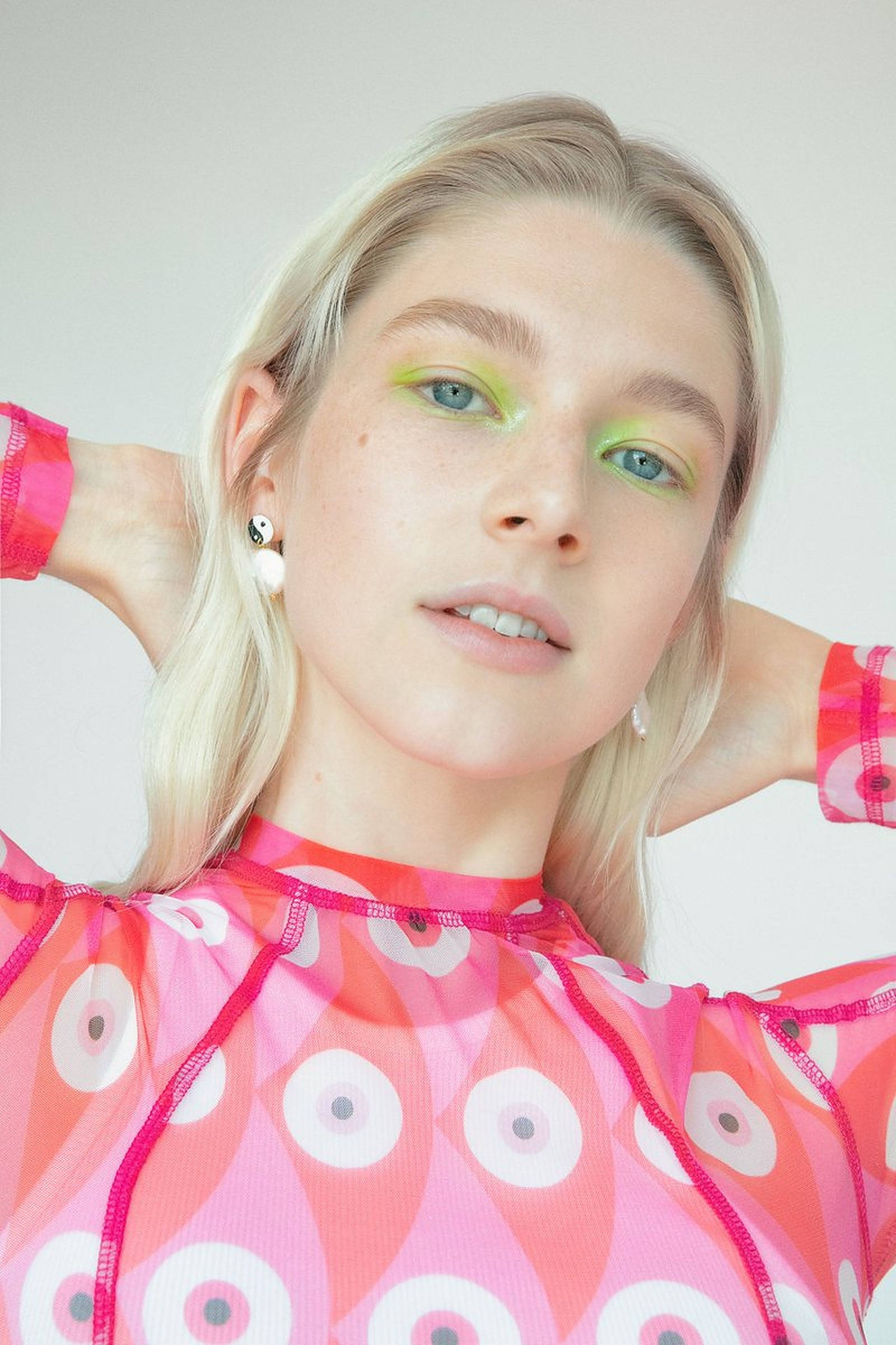Hunter Schafer 粉綠配色 壁紙 — 一位金髮女子塗上螢光綠色眼影，身穿帶有白色圓形圖案的透視粉紅色上衣，背景為純色。