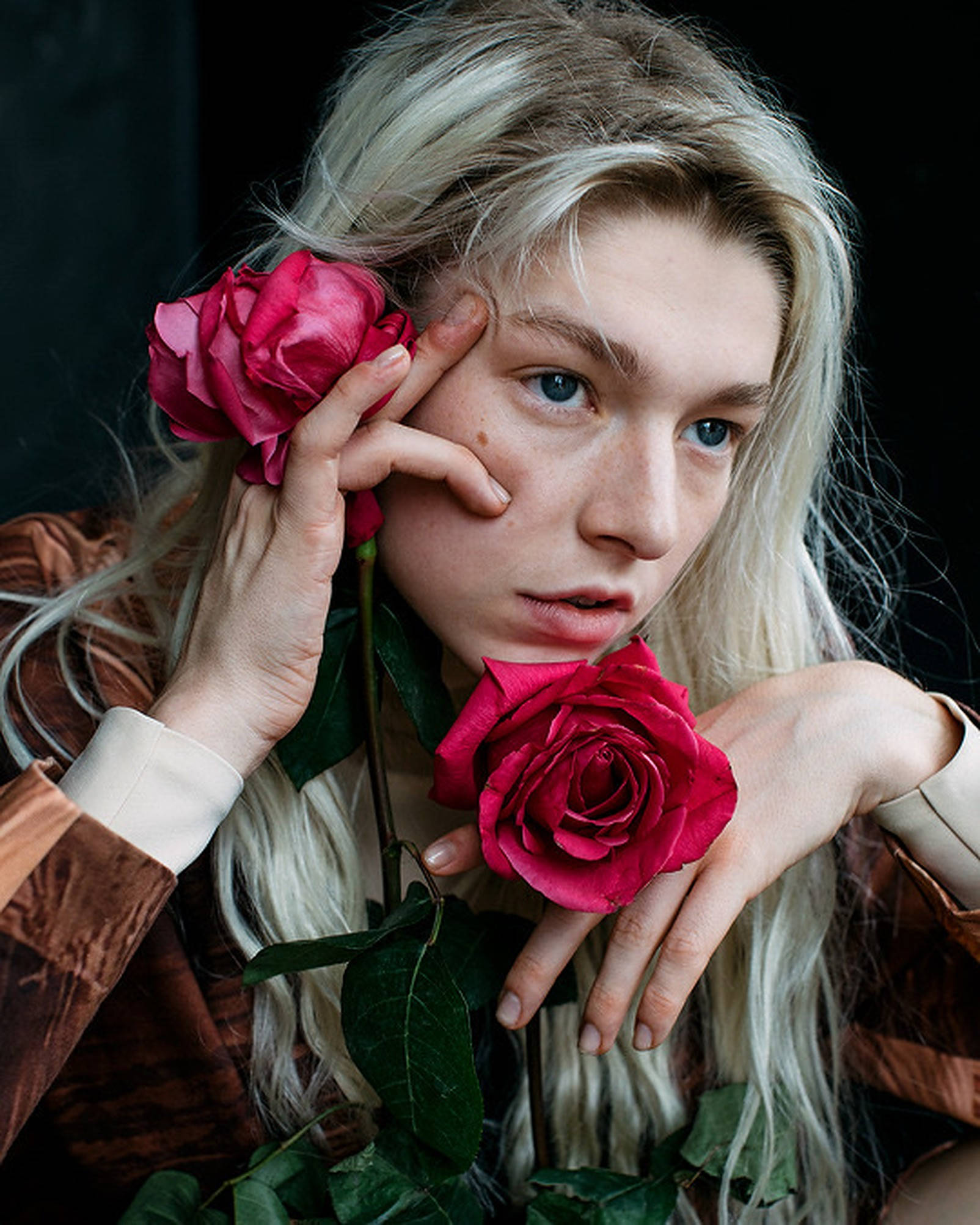 Hunter Schafer 紅玫瑰 壁紙 — 一位金髮年輕人在黑暗背景下手持鮮豔的紅玫瑰，營造出充滿氛圍的肖像。