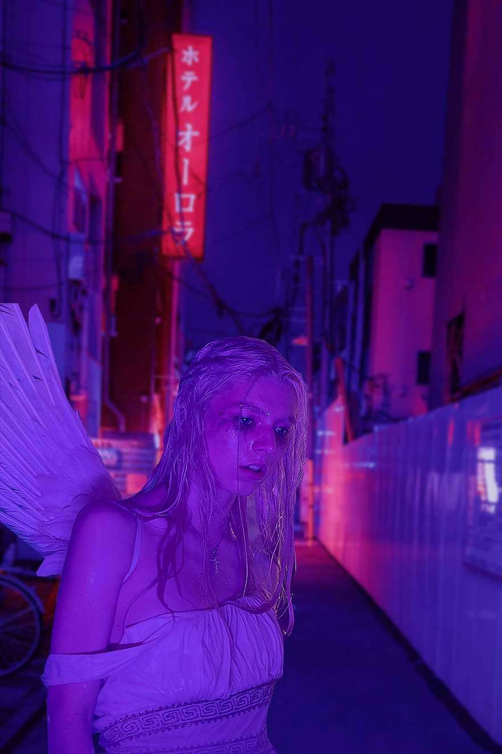 Hunter Schafer 紫色美學 壁紙 — 一位擁有天使翅膀的女性站在夜晚霓虹燈閃爍的小巷中，被發光招牌的紫色和粉紅色燈光籠罩。