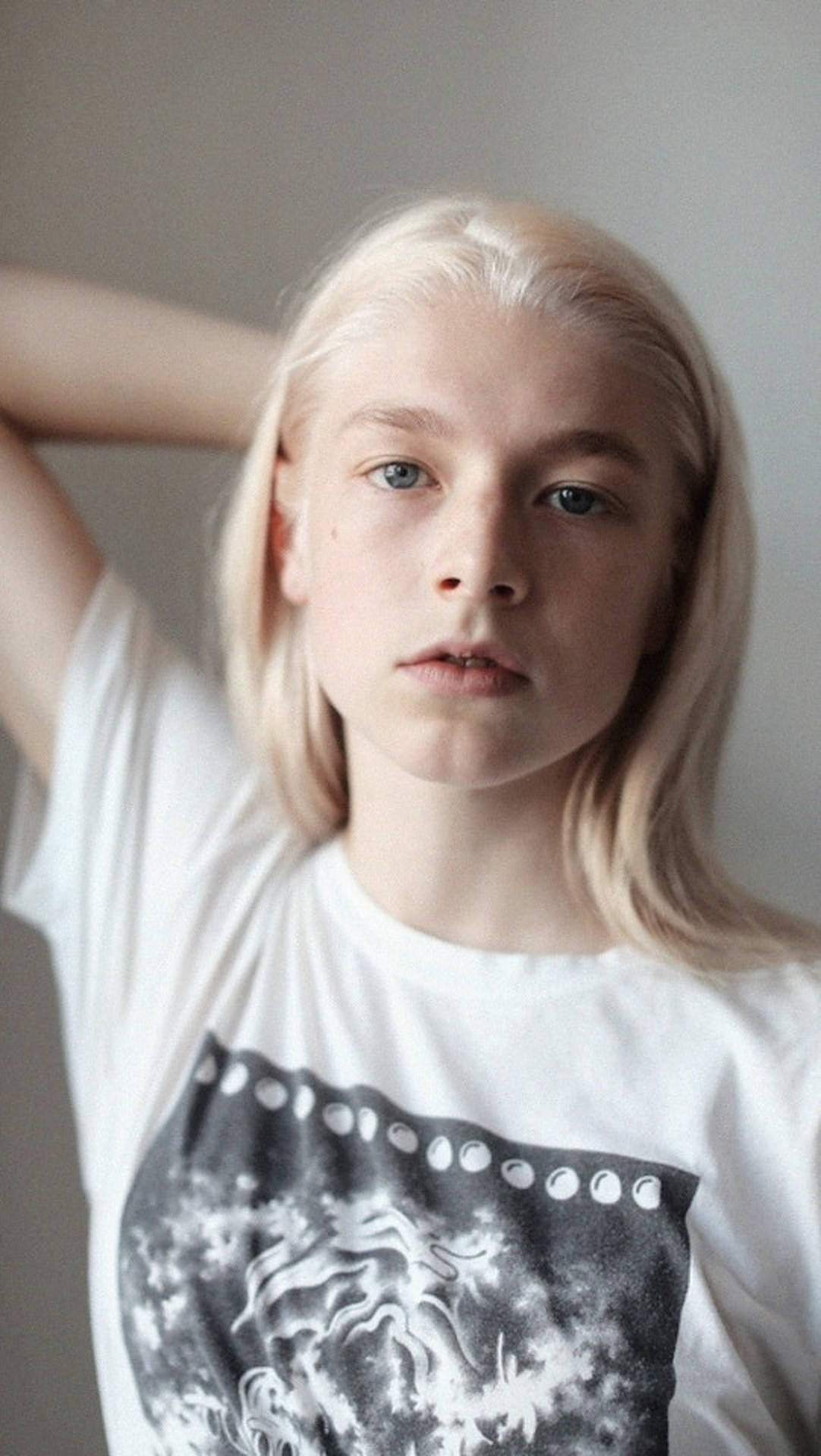 Hunter Schafer 自然妝容 壁紙 — 一位年輕人有著鉑金色頭髮和藍色眼睛，直接看著鏡頭，身穿帶有黑色圖案印花的白色T恤。