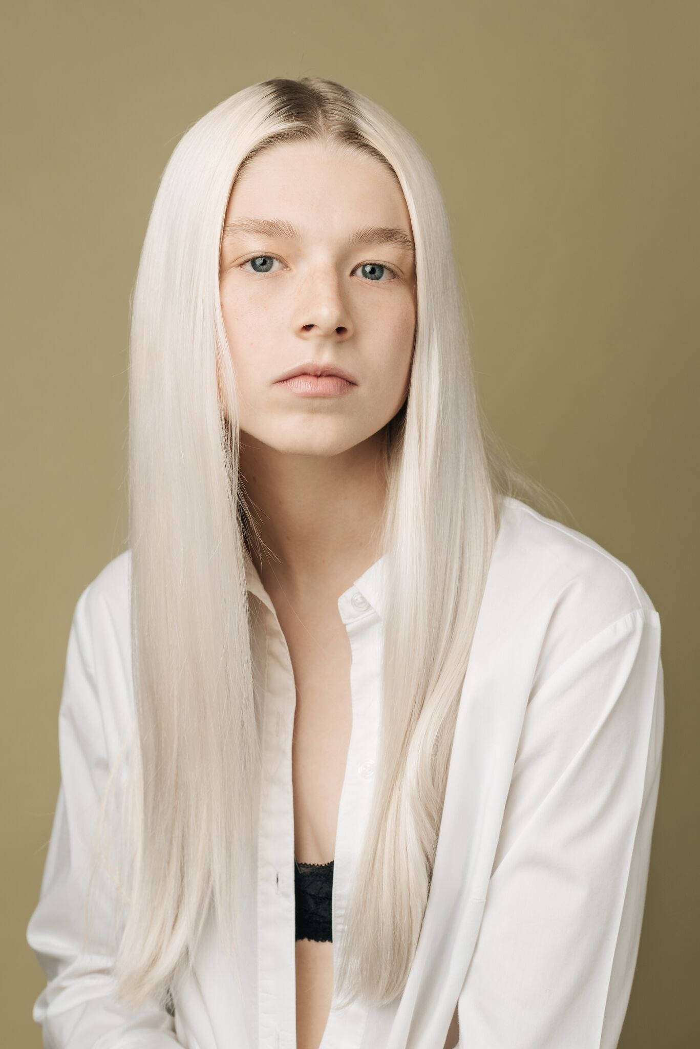 Hunter Schafer 銀髮造型 壁紙 — 一位年輕人有長白金髮和藍眼睛，穿著敞開的白襯衫，在柔和的橄欖綠背景前直視鏡頭。