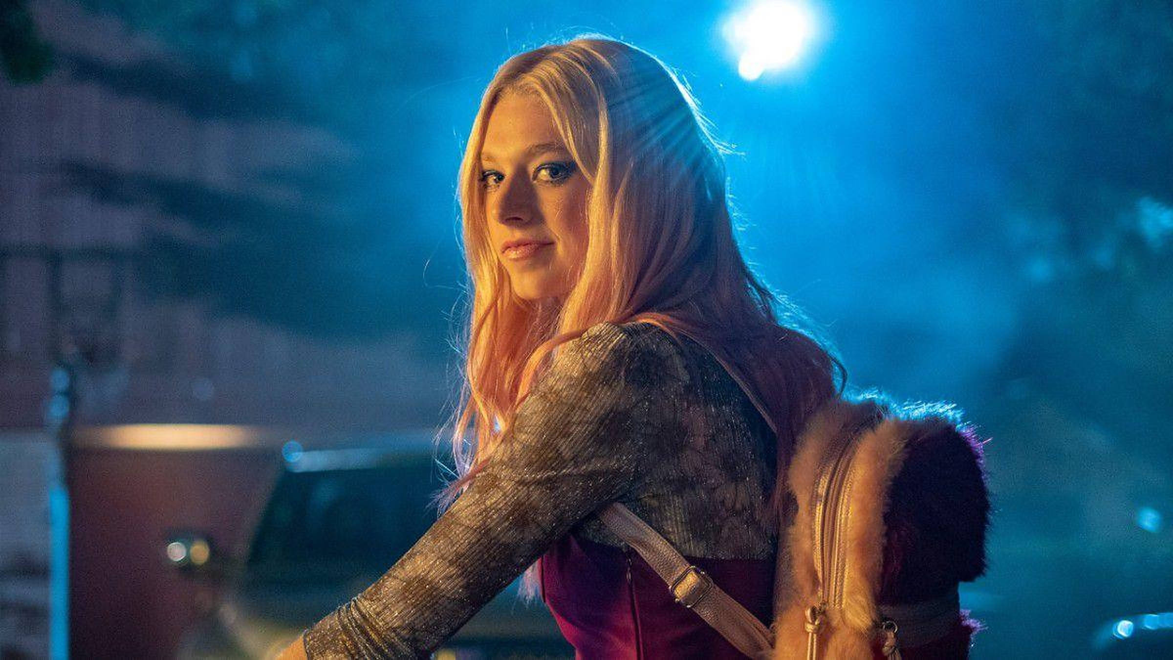 Hunter Schafer 飾 Jules 壁紙 — 一位年輕的長髮金髮女性在夜晚站在明亮的藍色聚光燈下，穿著質感上衣和毛邊背包。