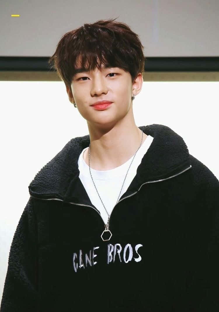 韓國藝人Hyunjin 時尚連帽衫 壁紙 — 一位黑髮、笑容溫和的年輕男子，在純色背景前穿著黑色抓絨連帽衫，內搭白色T恤。