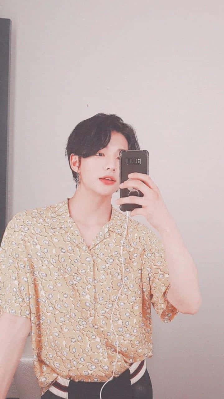 韓國藝人Hyunjin 柔焦自拍 壁紙 — 一位黑髮年輕男子穿著圖案黃色襯衫和白色耳機，在素牆前對著鏡子自拍。