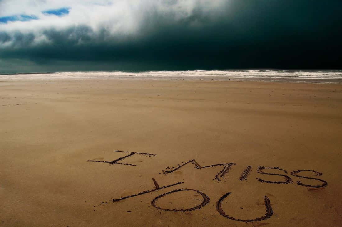 I Miss You 桌布 壁紙 — 「I MISS YOU」這幾個字刻在荒蕪海灘的濕沙上，上方籠罩著一片迫近的黑暗暴風雲。