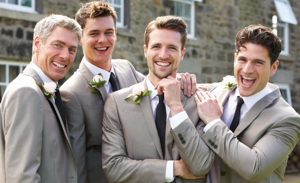 婚禮前與伴郎們的歡樂時光！（由左至右）Ian、Howard、Dave 和 Jason——最棒的伴郎團！ 壁紙 — 四位身穿灰色西裝的快樂伴郎，在石建築背景下，佩戴白色胸花一起大笑。