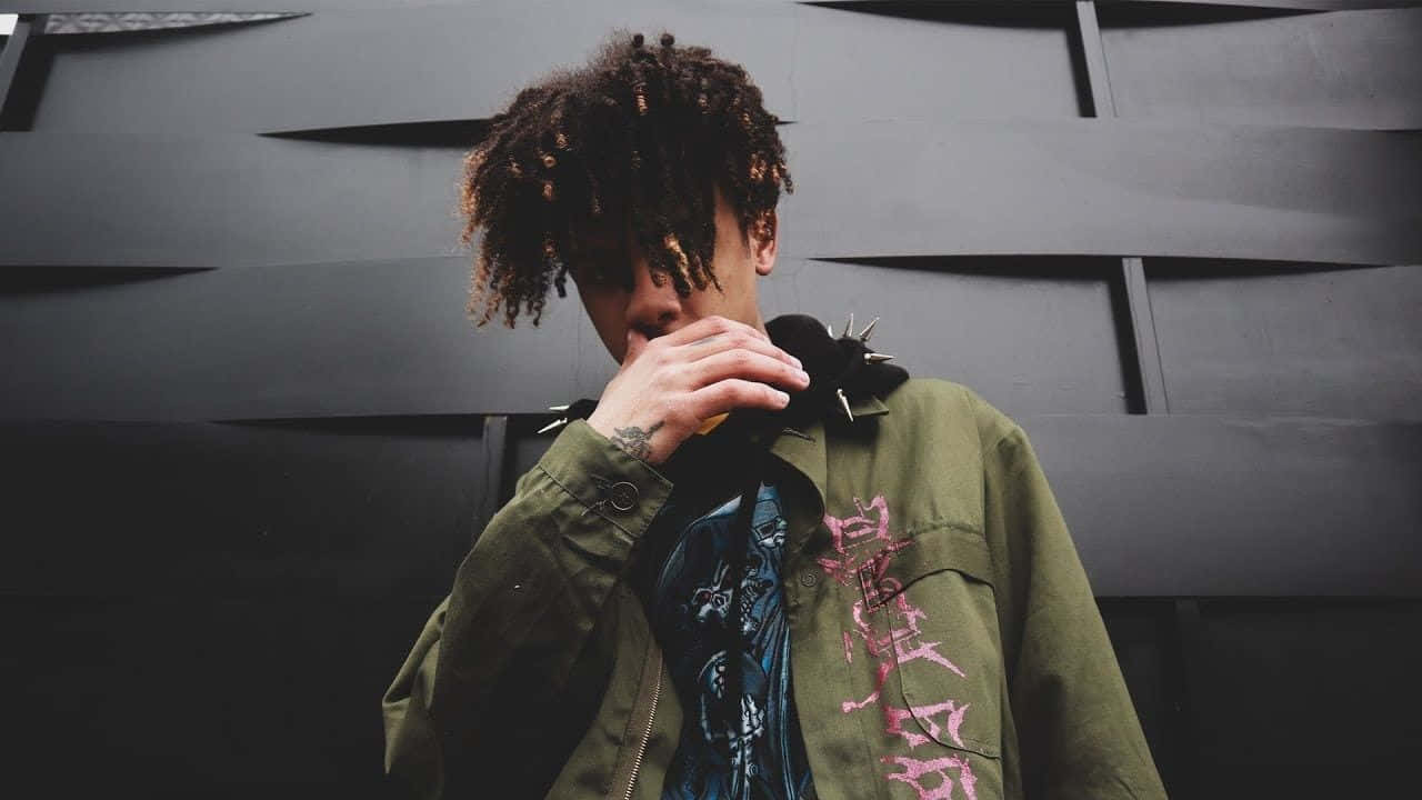 Iann Dior 姿態照 壁紙 — 一個捲髮、身穿橄欖色外套的人站在深色幾何牆前，手靠近嘴邊。