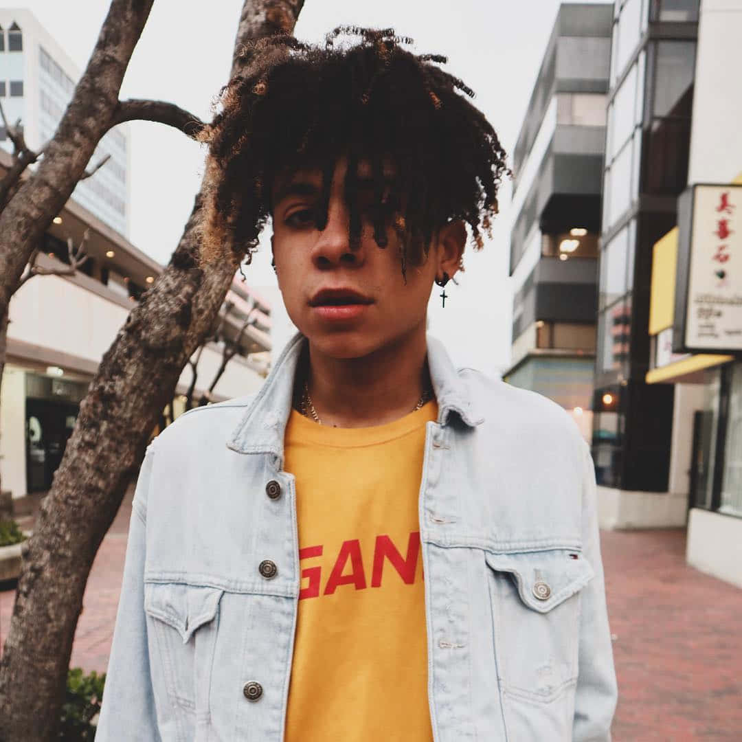 Iann Dior 戶外漫步 壁紙 — 一位年輕人有曲髮，身穿牛仔褸，站在城市行人路上，旁邊有樹，背景是現代建築。