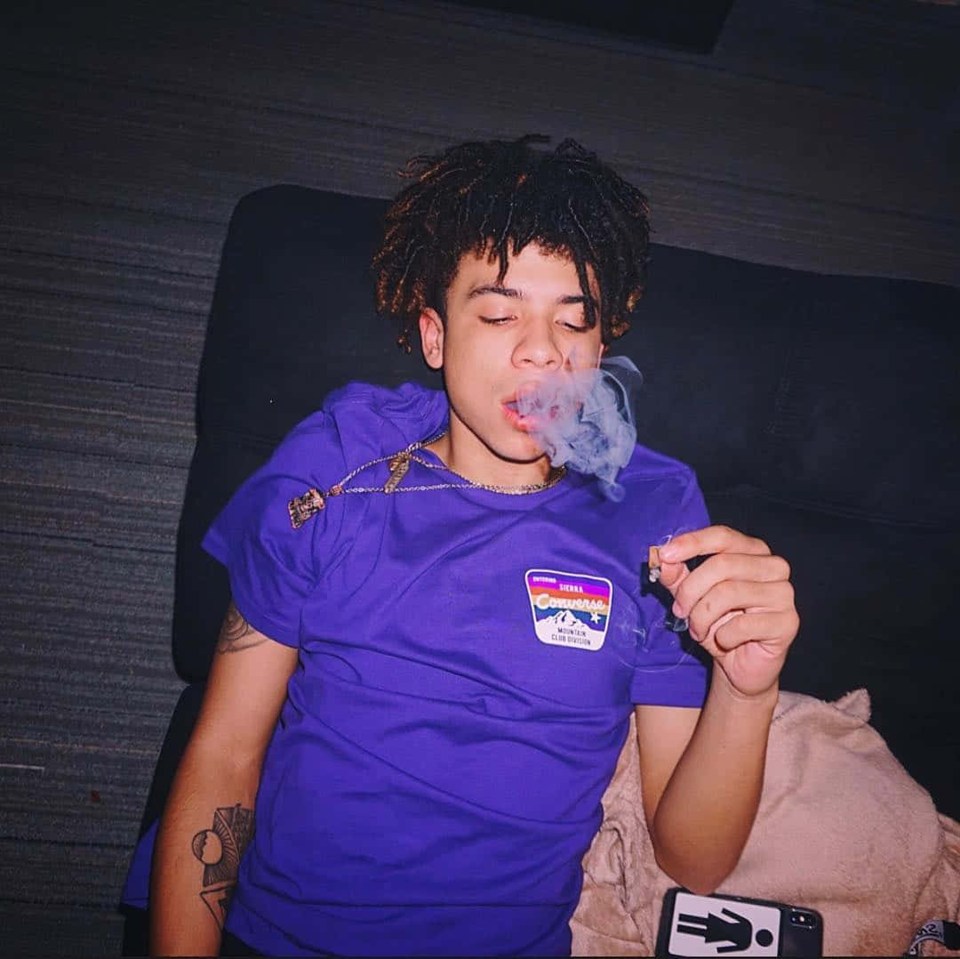 Iann Dior 躺著抽菸 壁紙 — 一位年輕的捲髮人士在昏暗的房間裡穿著紫色襯衫，躺在深色沙發上呼出煙霧。