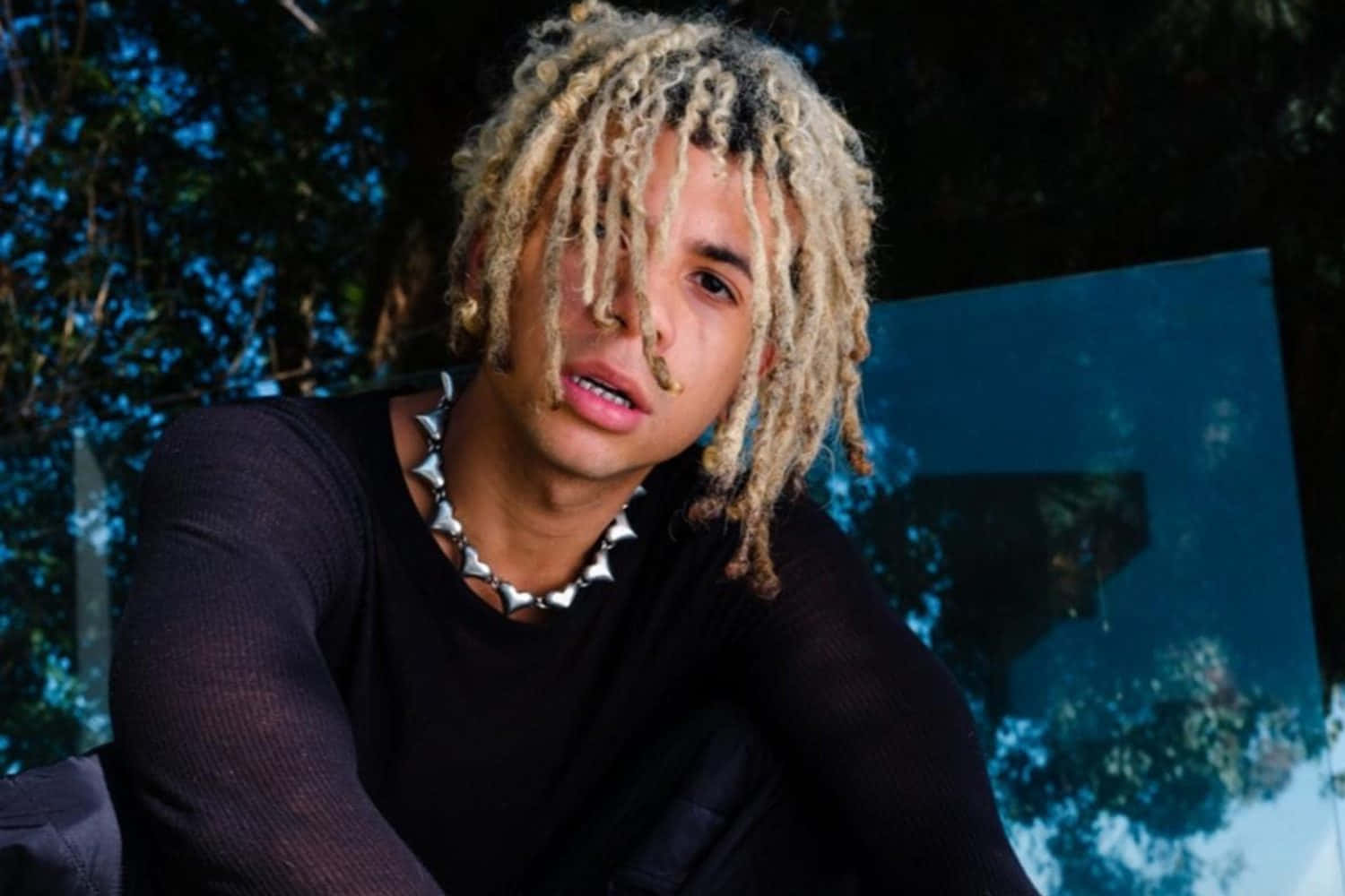 Iann Dior 金髮辮髮 壁紙 — 一位金髮辮子、戴著尖刺頸鏈的年輕男子，在深藍色抽象背景前凝視。