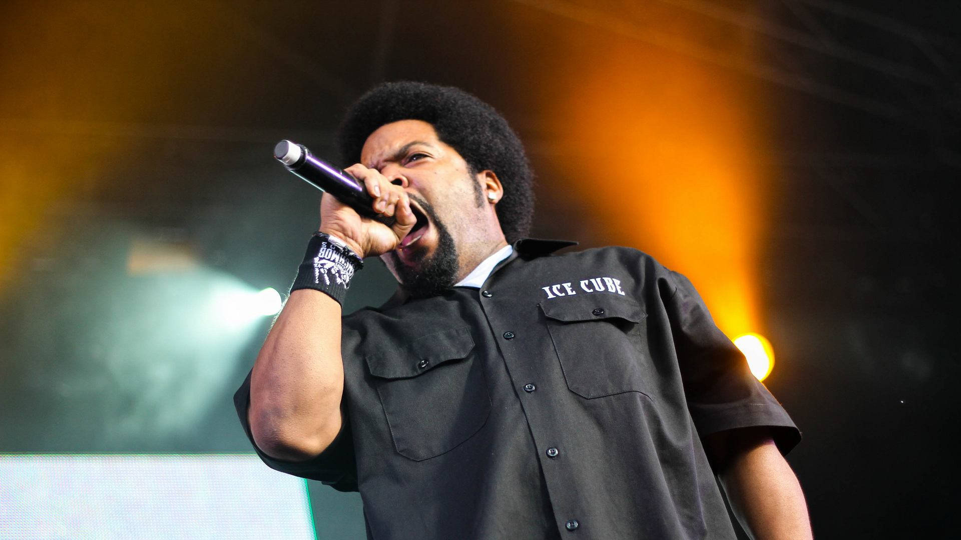 Ice Cube 在舞台上表演 壁紙 — 饒舌歌手 Ice Cube 在溫暖的橙色和綠色舞台燈光下，熱情地握著麥克風表演。