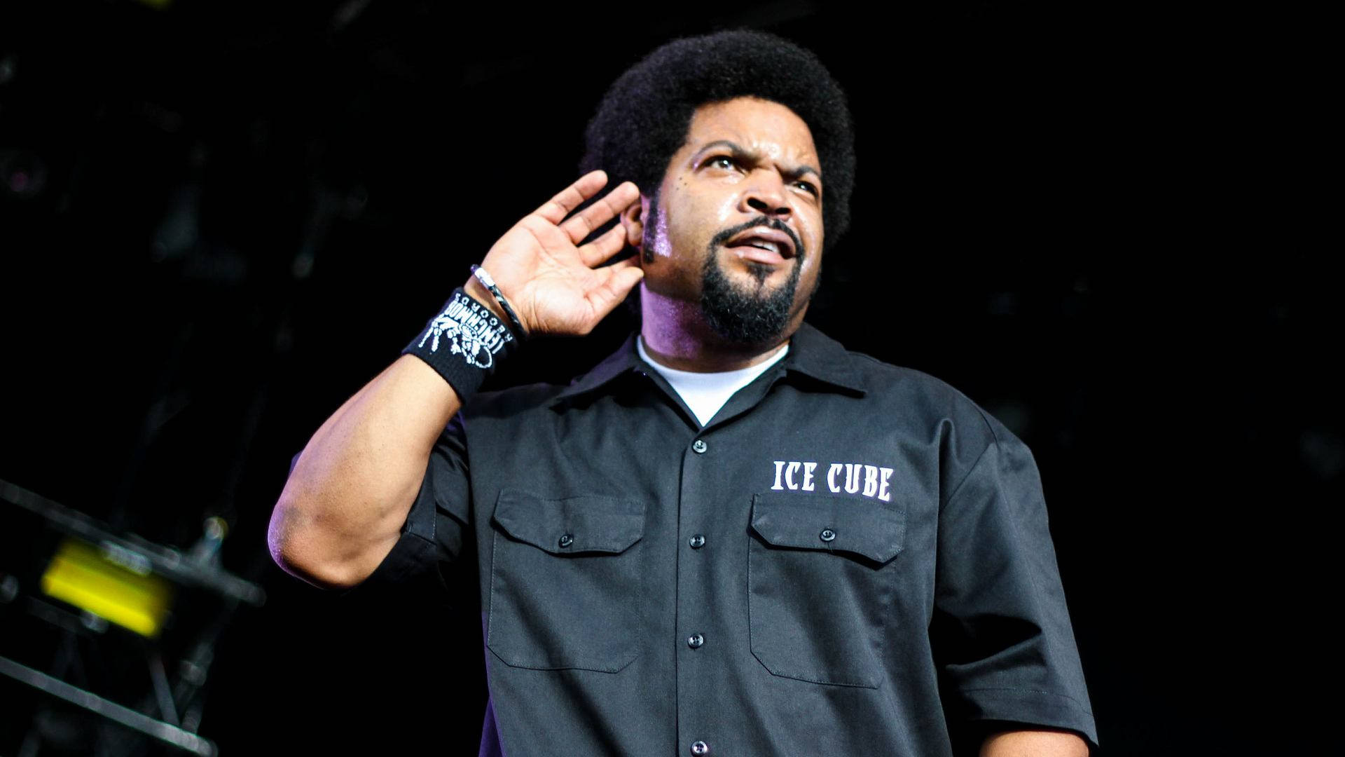 Ice Cube 手貼耳 壁紙 — Ice Cube 在舞台上表演，手呈杯狀放在耳邊，在明亮的舞台燈光下，背景黑暗。