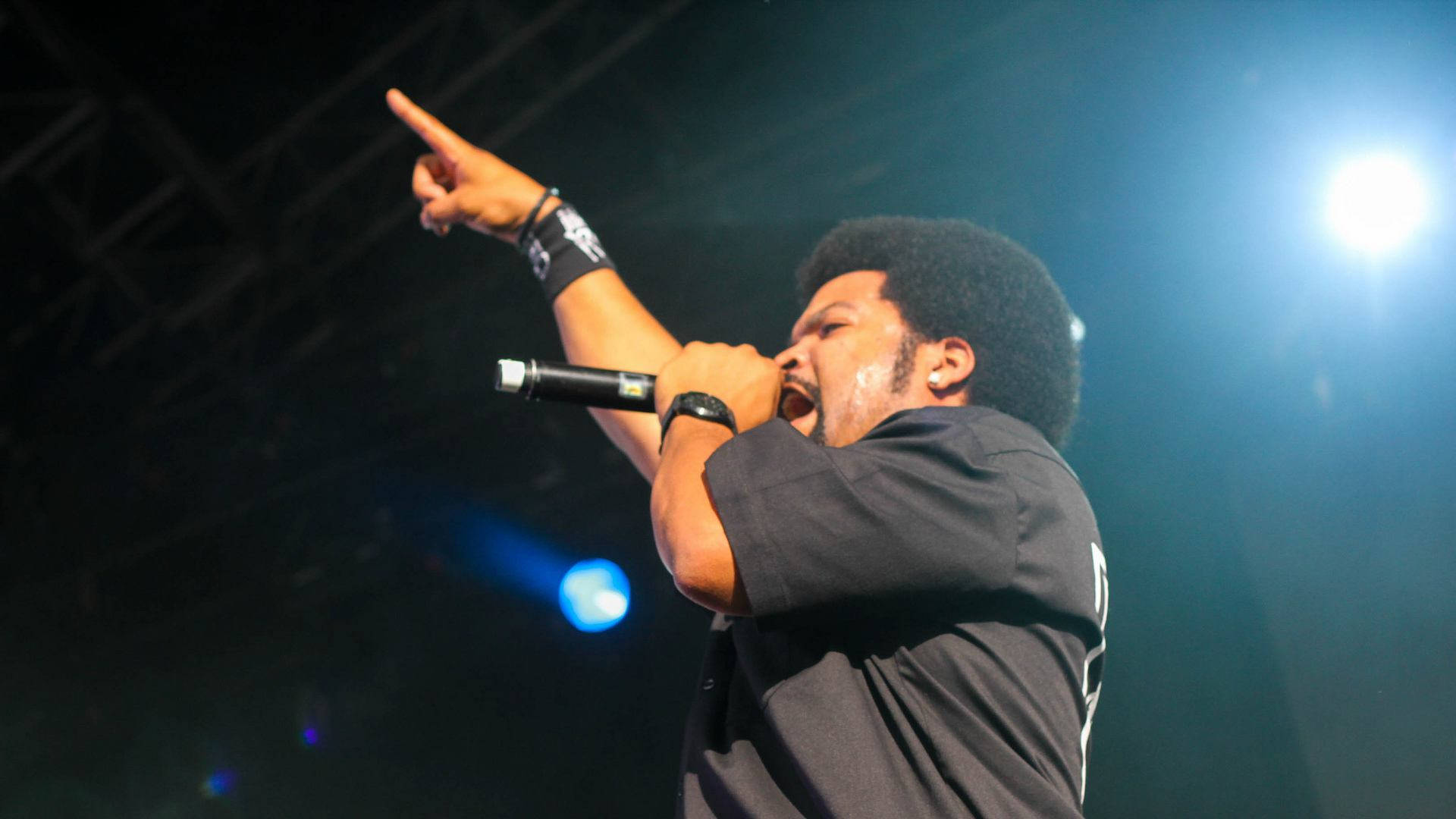 Ice Cube 表演中 壁紙 — 一位留著非洲髮型的男性表演者在明亮的舞台燈光下熱情地對著麥克風唱歌，同時向上指著。