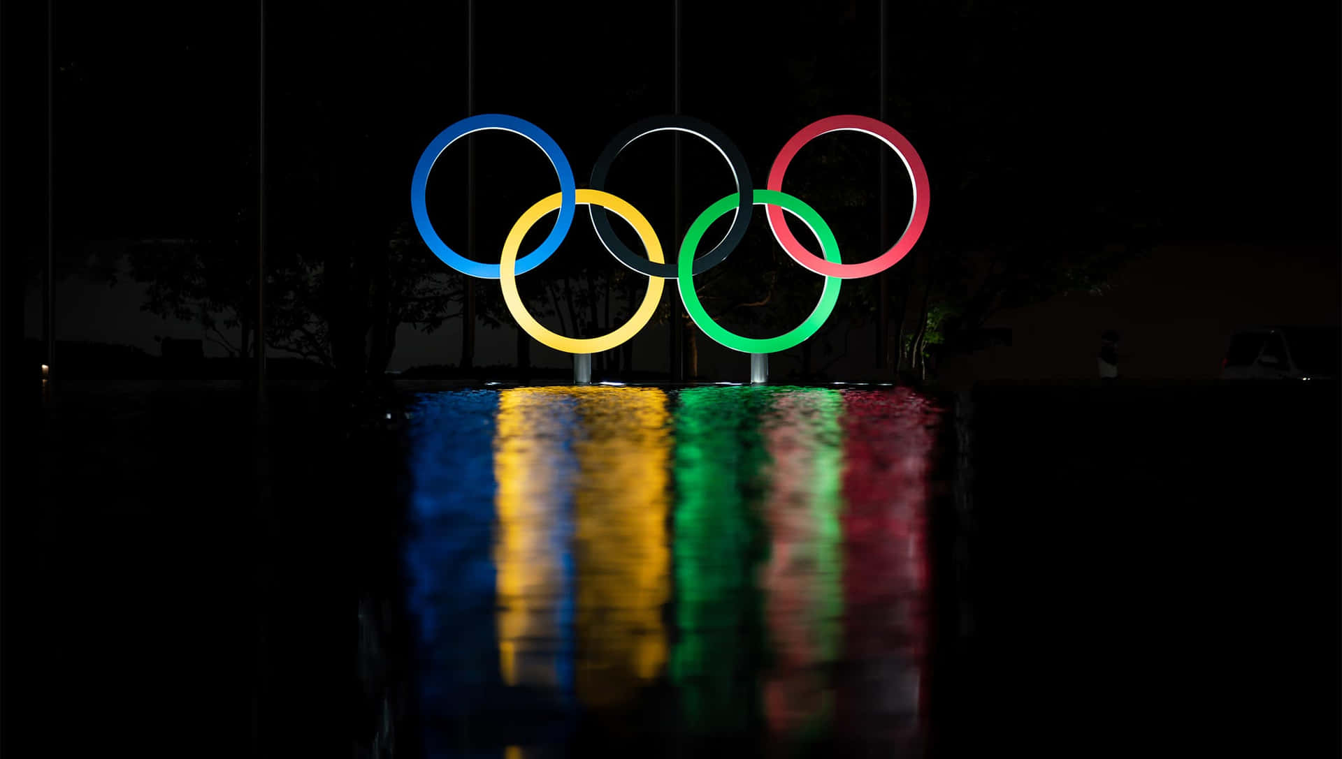 夜晚點亮的奧運五環，在潮濕地面上有彩色倒影。