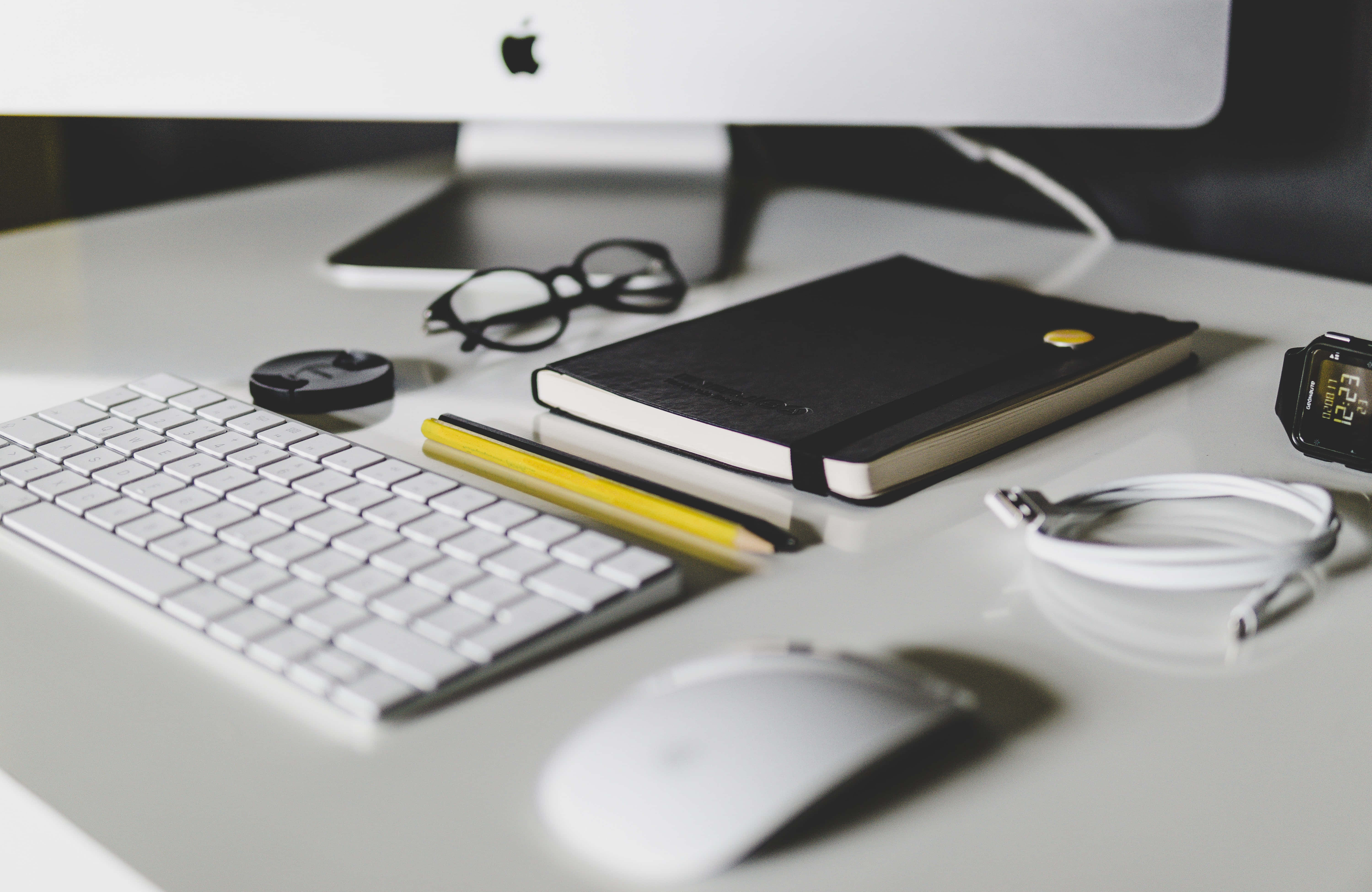 蘋果 iMac 電腦螢幕背景 壁紙 — 簡約白色桌面設置，配有鍵盤、滑鼠、筆記本和眼鏡，在柔和日光下。