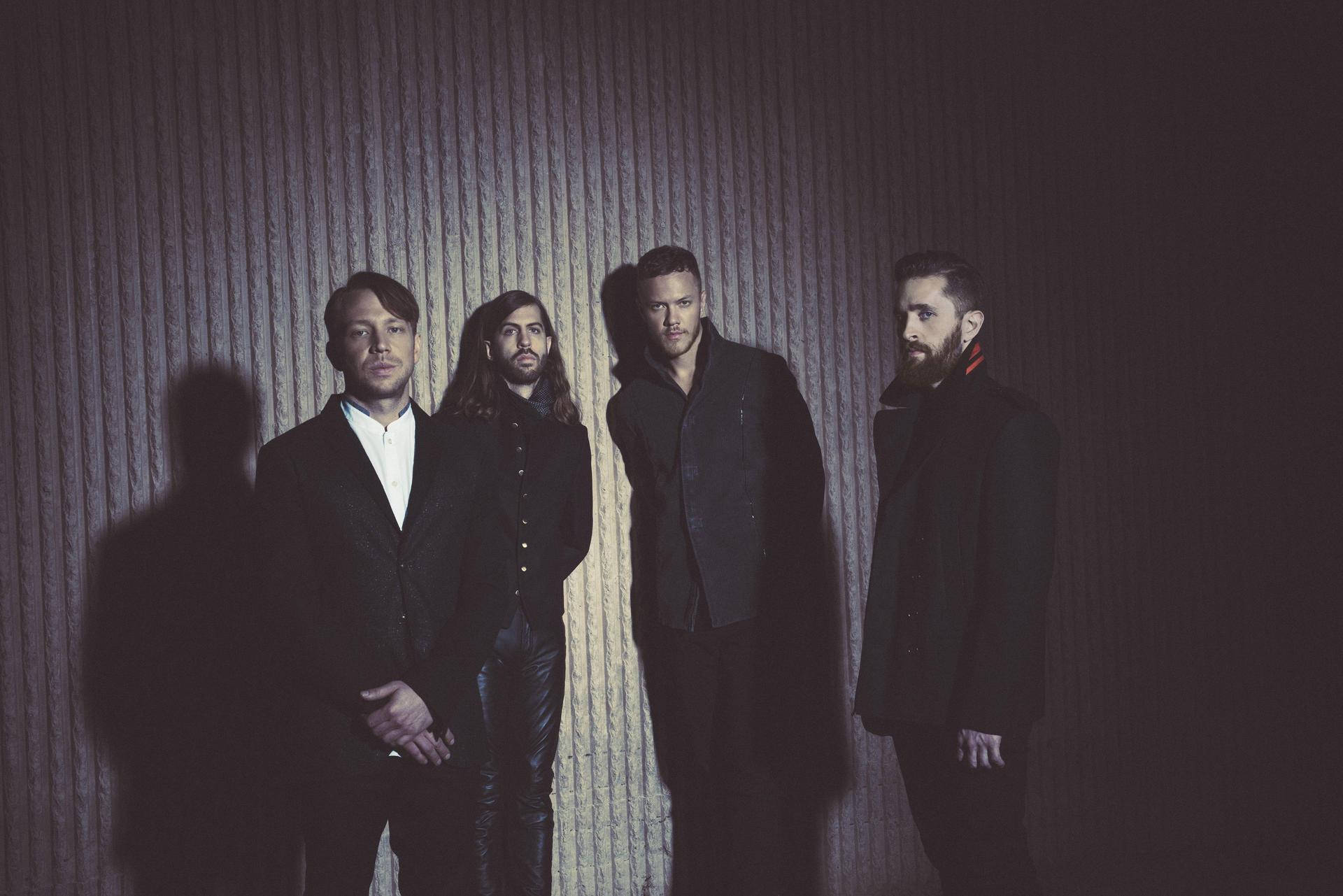 Imagine Dragons 2015專輯拍攝現場 壁紙 — 四個身穿深色西裝的男人在戲劇性的聚光燈下，對著有紋理的牆壁莊嚴地站立，投射出長長的影子。