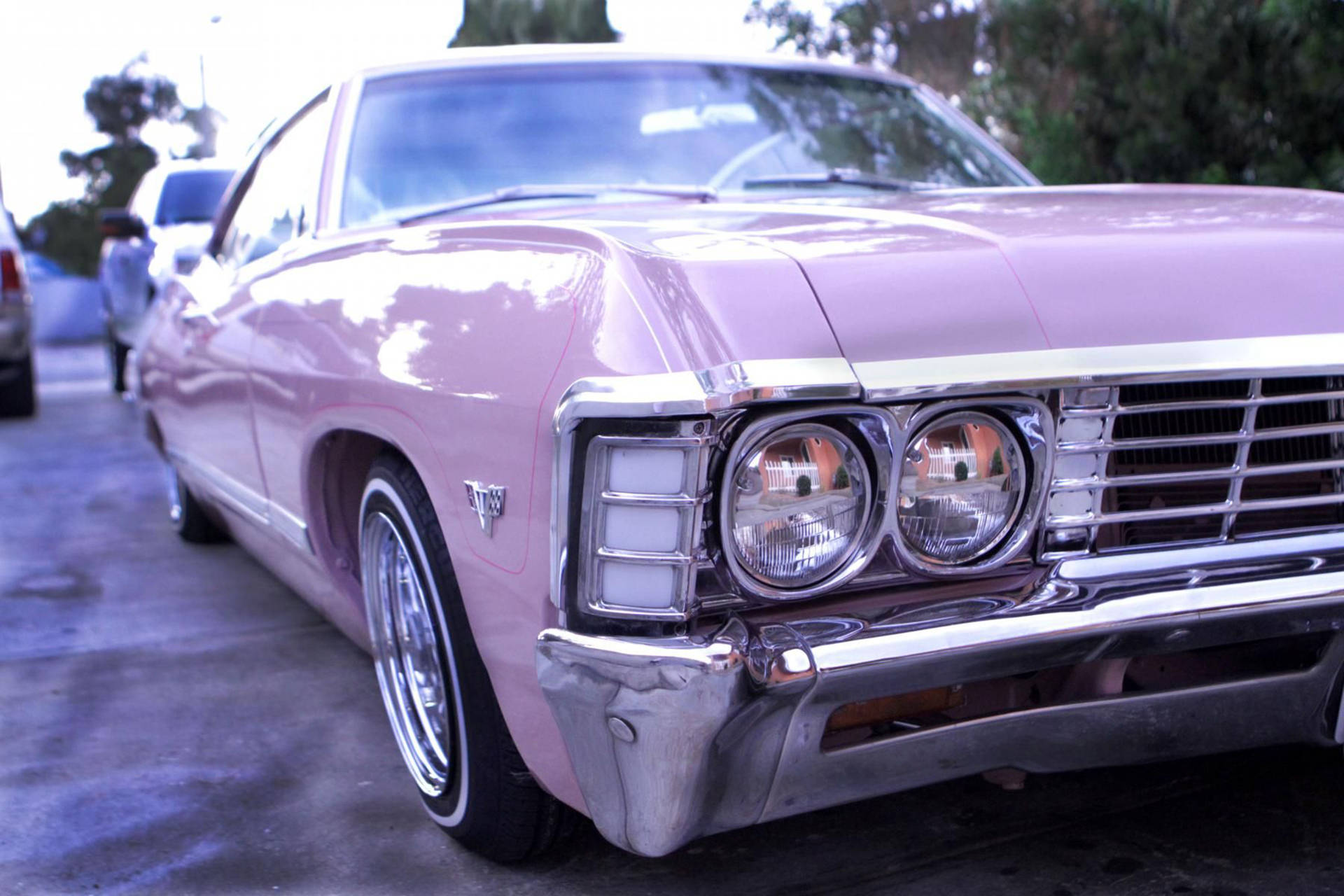 紫色雪佛蘭Impala 1967 壁紙 — 一輛復古粉紅色肌肉車，帶有鍍鉻細節，在停車場的柔和日光下閃閃發光。