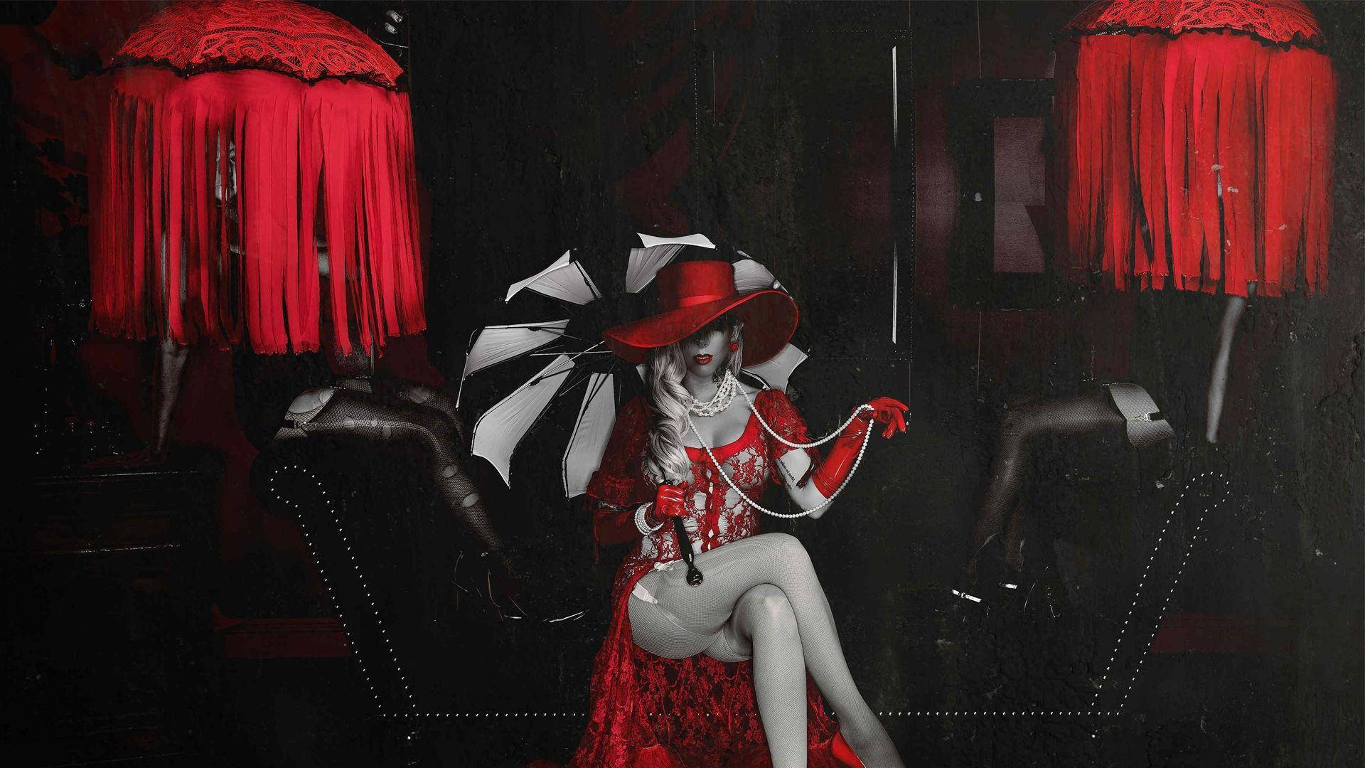 In This Moment《Bloody Creature》藝術圖 壁紙 — 一位身穿紅色蕾絲連衣裙和帽子的女士優雅地坐在黑色皮椅上，兩側是紅色流蘇燈，房間昏暗而富有氣氛。