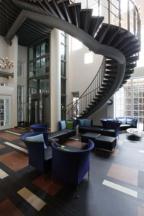 Inntel Hotel Zaandam 的樓梯 壁紙 — 一個宏偉的現代大堂設有華麗的螺旋樓梯，配有深色金屬踏板和玻璃欄杆，周圍環繞著深藍色休閒椅和圖案瓷磚地板。