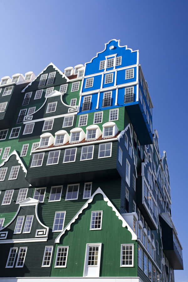 Inntel Hotel Zaandam 的角落藍屋 壁紙 — 一座引人注目的現代建築，設計成類似堆疊的荷蘭山牆房屋，以綠色、藍色和白色呈現，背景是清澈的藍天。