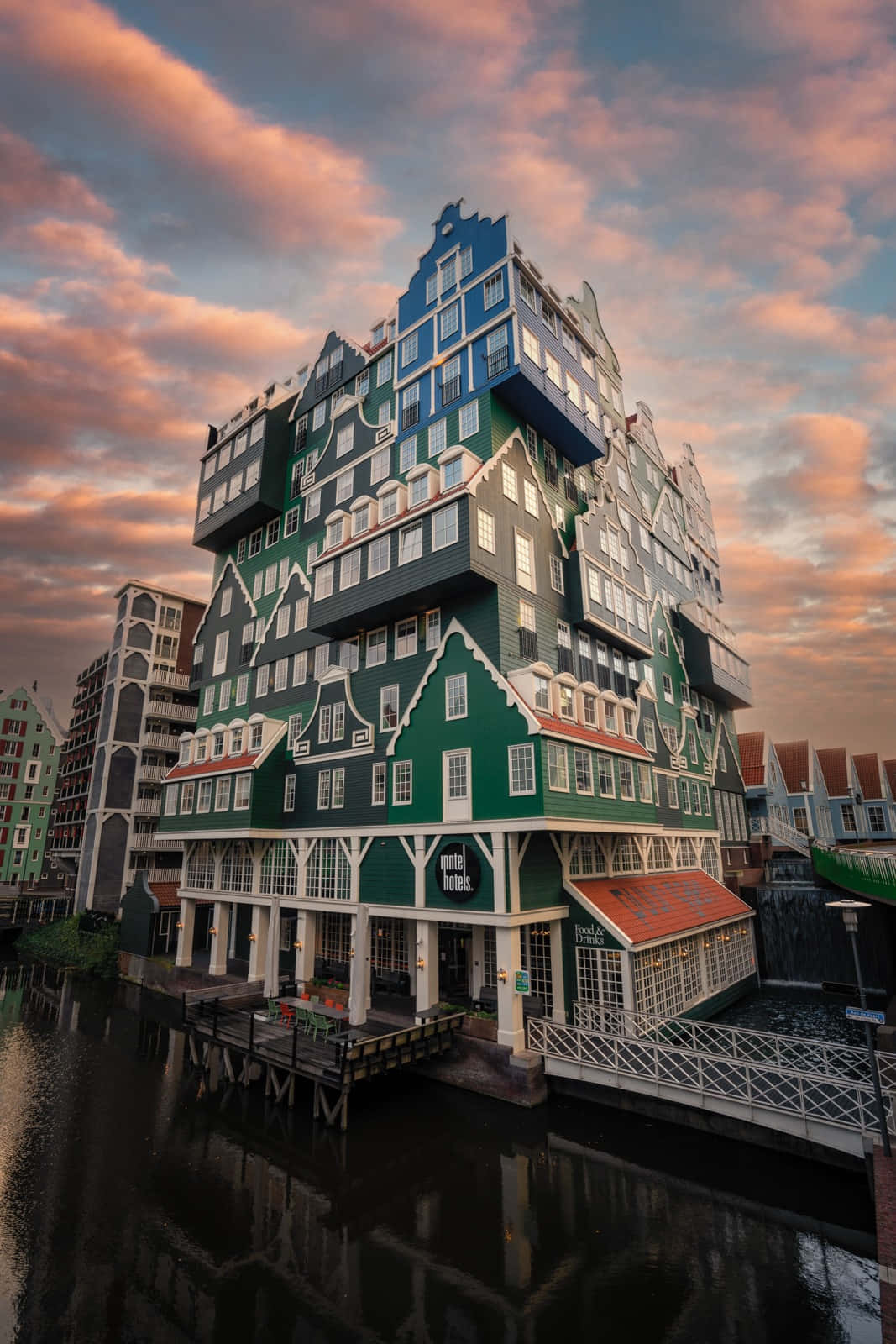 Inntel Hotel Zaandam 在絢麗天空下 壁紙 — 一座引人注目的綠色和藍色現代酒店，像荷蘭山牆房屋一樣堆疊，在戲劇性的日落天空下倒映在平靜的運河水中。