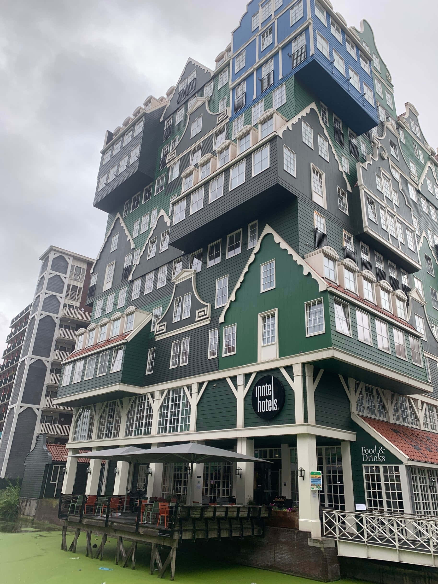 Inntel Hotel Zaandam 雲層天空 壁紙 — 一座引人注目的綠色和藍色階梯式山牆酒店建築，帶有白色飾邊，矗立在綠色運河之上，天空陰沉。