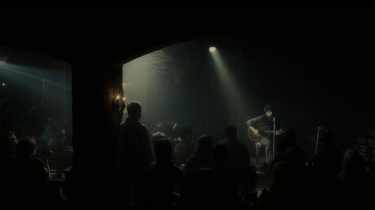 《Inside Llewyn Davis》舞台演出 壁紙 — 一位孤獨的結他手在昏暗的親密場地中，於單一聚光燈下表演，觀眾以剪影呈現。