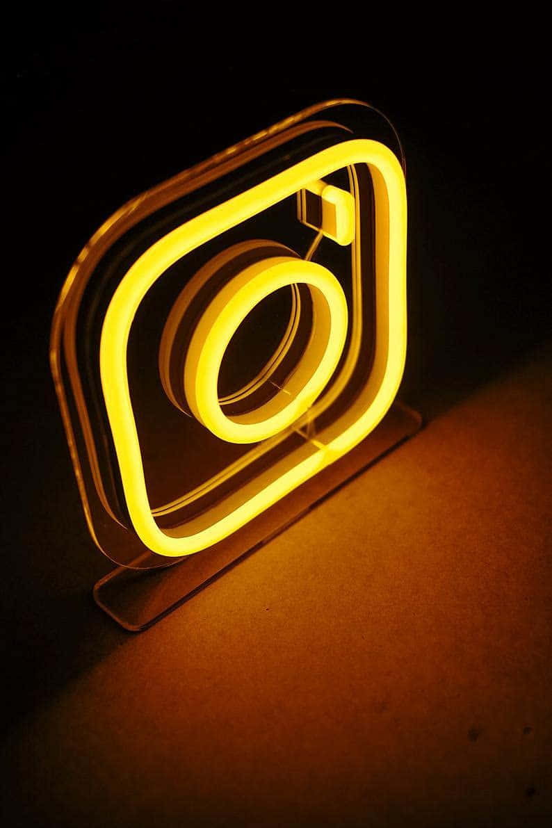 Instagram標誌燈光圖 壁紙 — 一個發光黃色霓虹燈牌，形狀像Instagram標誌，在黑暗表面上投射出溫暖的光芒。