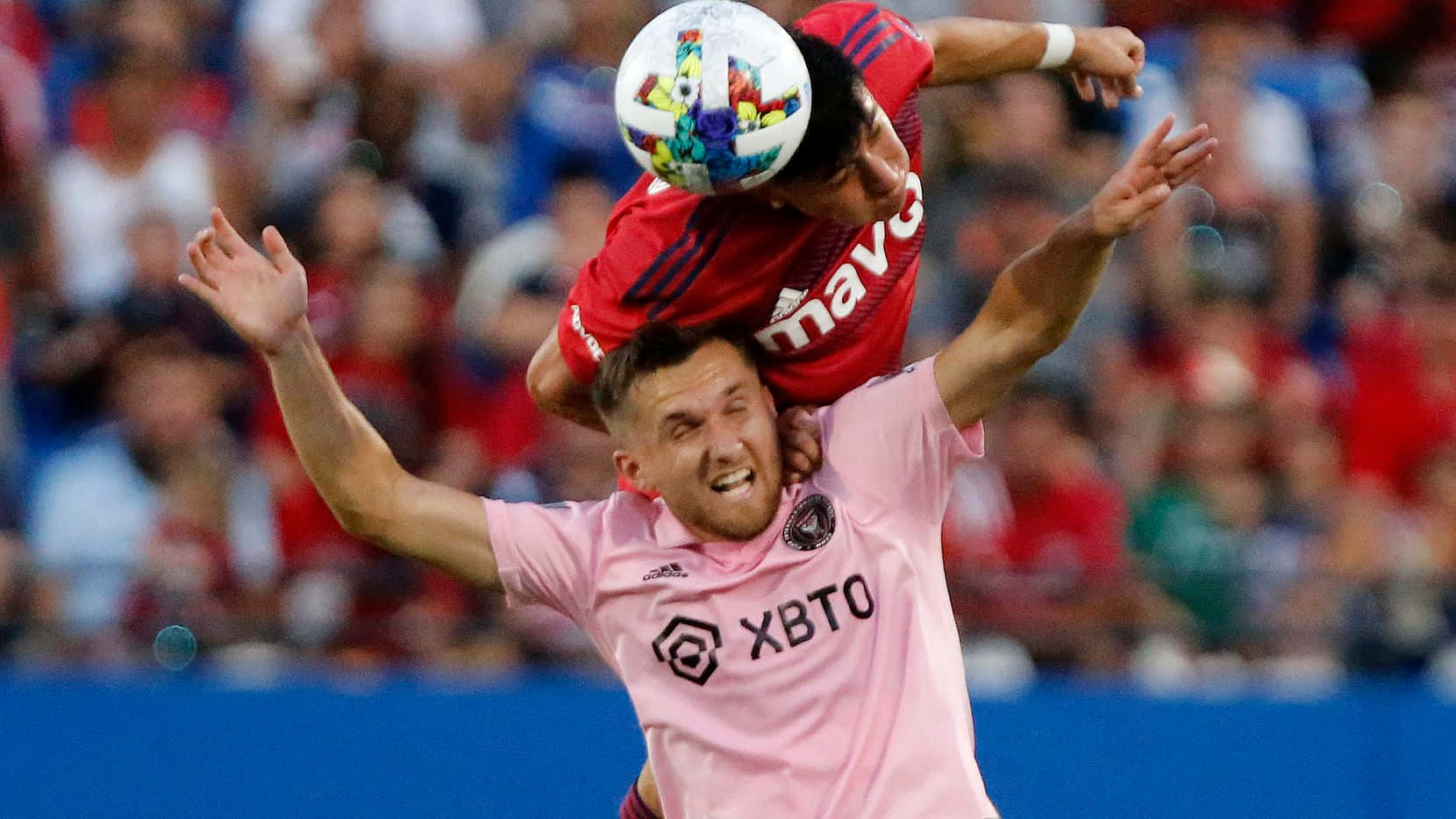 Inter Miami FC 中場 Indiana Vassilev 與 FC Dallas 球員 Marco Farfan 壁紙 — 兩名足球運動員在半空中跳起，爭奪一個色彩繽紛的球，背景是模糊的體育場人群。