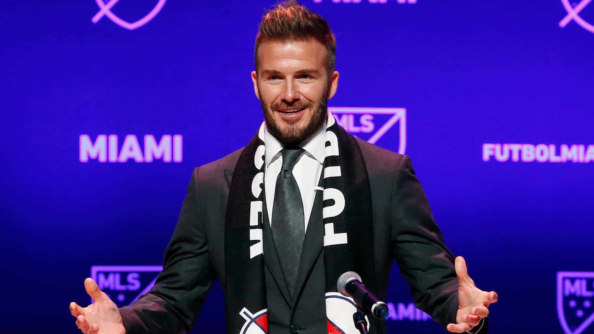 Inter Miami FC 主席兼擁有者大衛·貝克漢 中景肖像 壁紙 — 大衛碧咸在鮮藍色背景下出席記者會，溫暖微笑，戴著邁阿密FC圍巾。