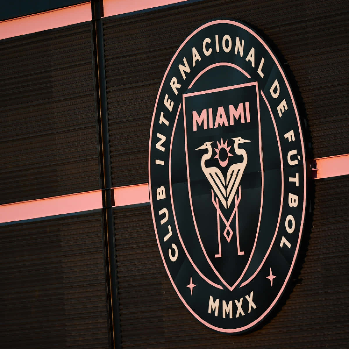 Inter Miami FC 官方隊徽印章側面視角 壁紙 — 一個發光的粉紅色和黑色 Club Internacional de Fútbol Miami 標誌安裝在帶有水平燈條的深色紋理牆壁上。