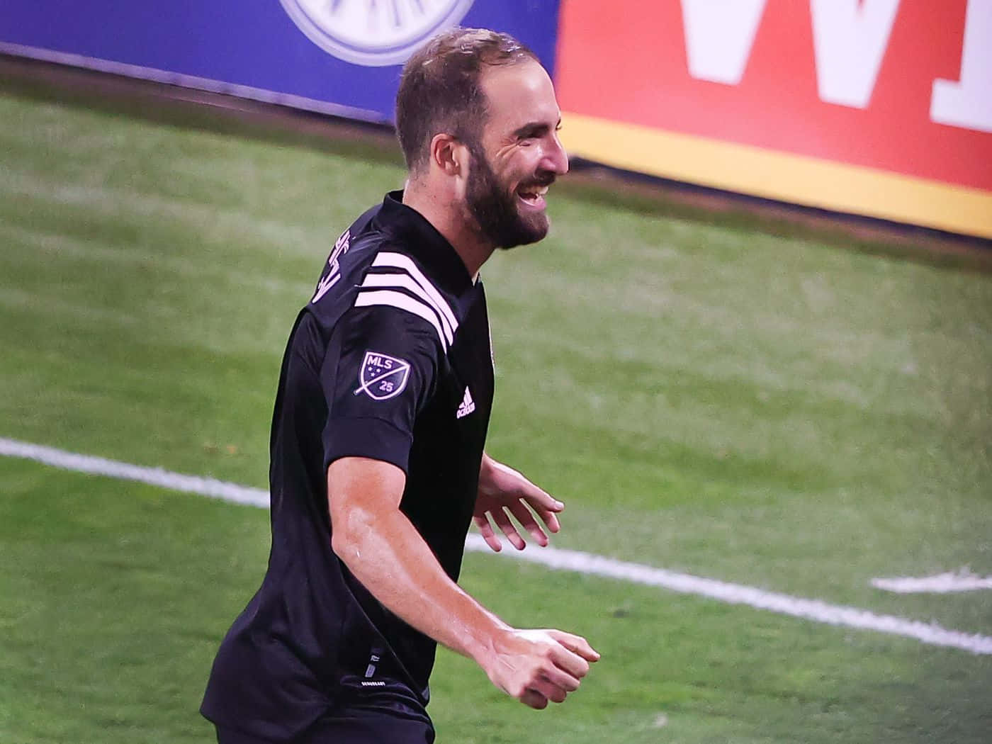 Inter Miami FC 球員 Gonzalo Higuain 側面賽場照 壁紙 — 一名身穿黑色球衣的足球運動員在體育場燈光下，笑著跑過充滿活力的綠色球場。