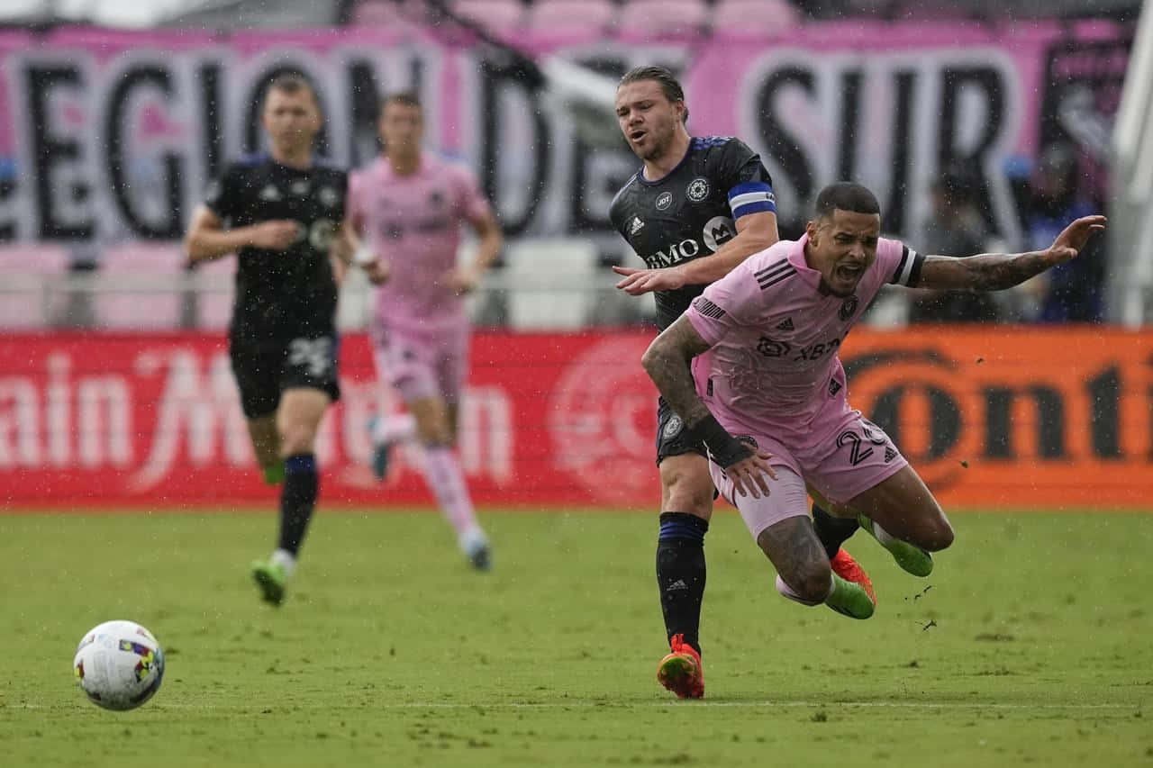 Inter Miami FC 與 FC Montreal 場上激烈對決 壁紙 — 一名身穿粉紅色球衣的足球員在雨中比賽中撲向球，而一名身穿黑色的後衛正在逼近。
